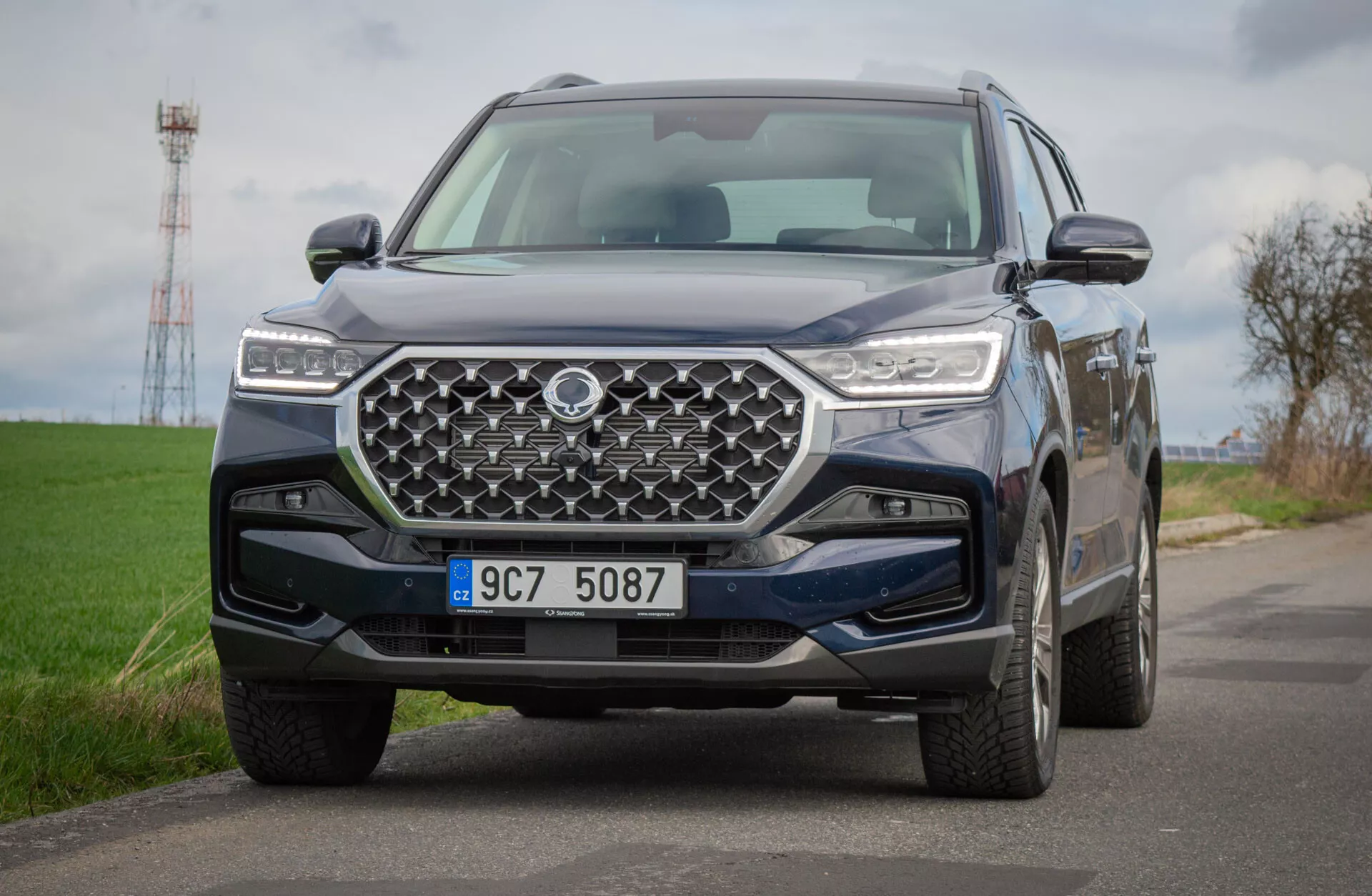 SUV SsangYong Rexton 2021 na wiejskiej drodze, ukazujący szczegółowy widok jego przedniego projektu z geometryczną kratką i reflektorami LED, odzwierciedlający zaawansowaną technologię i możliwości terenowe