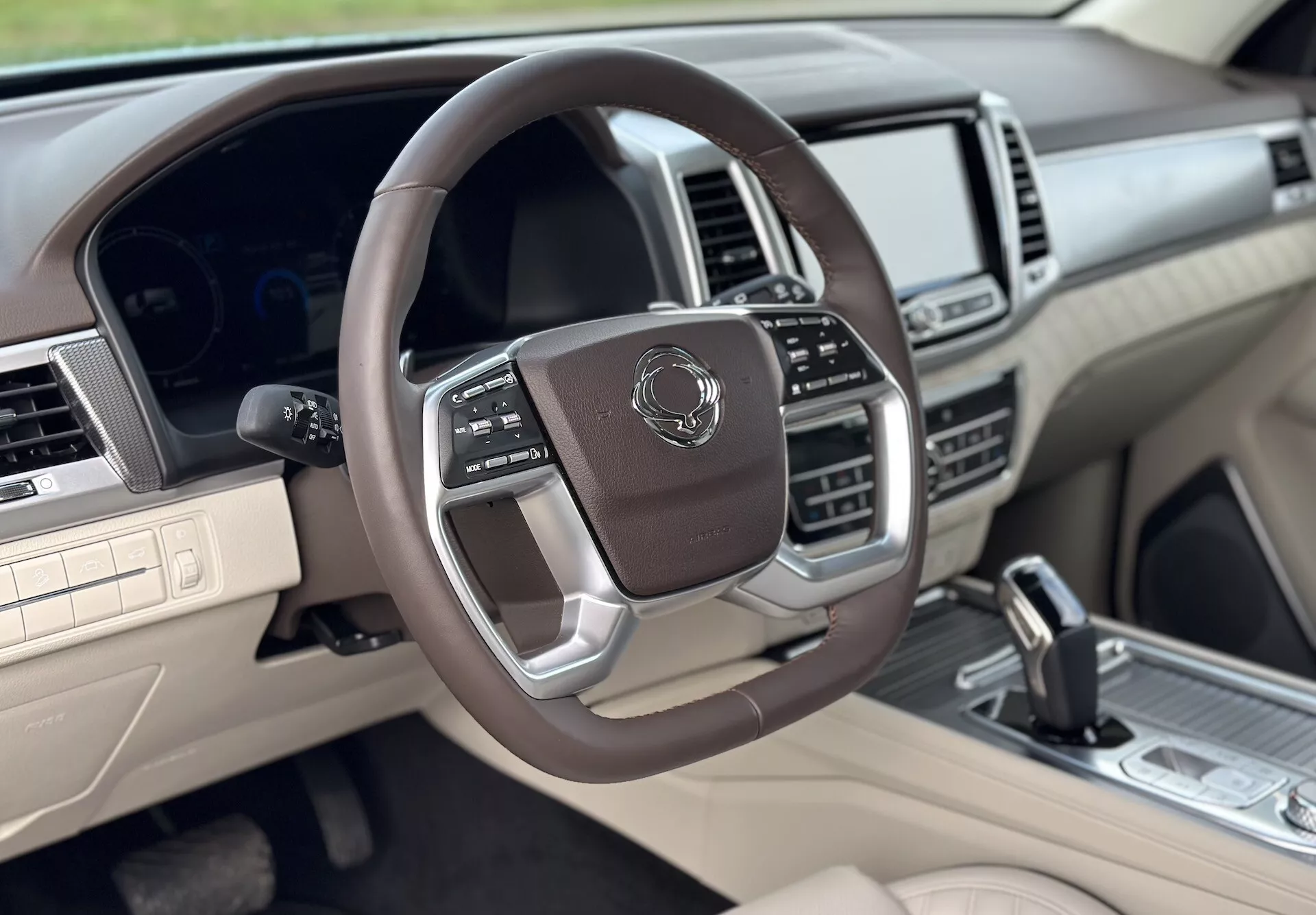 Luksusowe wnętrze SsangYong Rexton 2021 z perspektywy miejsca pasażera przedniego, prezentujące wielofunkcyjną kierownicę owiniętą skórą z metalicznymi akcentami, cyfrowy zestaw wskaźników, zaawansowany system infotainment i dwukolorową kolorystykę beżowo-ciemną.