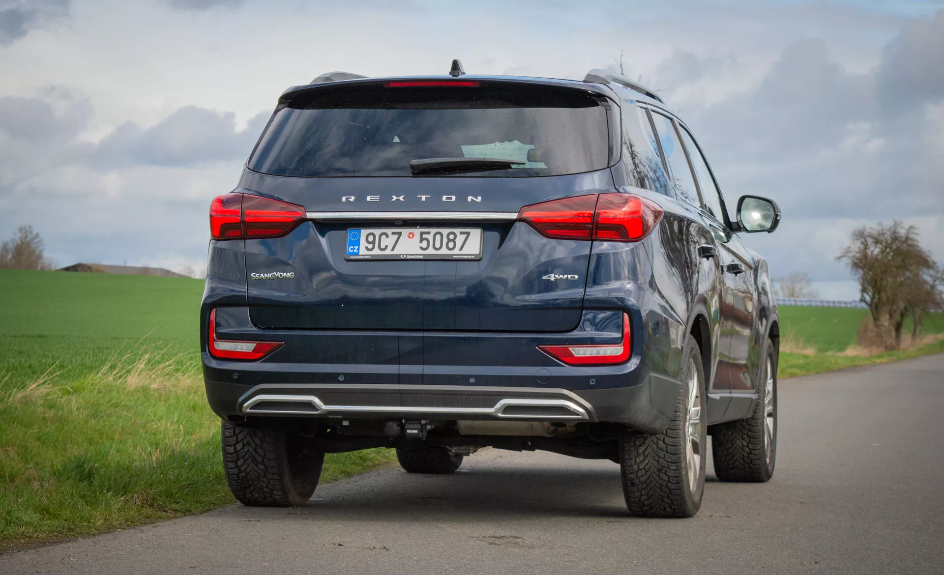 Tylny widok niebieskiego SUV-a SsangYong Rexton 2021 zaparkowanego na wiejskiej drodze dwupasmowej, podkreślający jego cechy konstrukcyjne, takie jak tylne światła, tylna szyba, rury wydechowe i technologia bezpieczeństwa, w tym kamera cofania i czujniki parkowania, pod pochmurnym niebem.