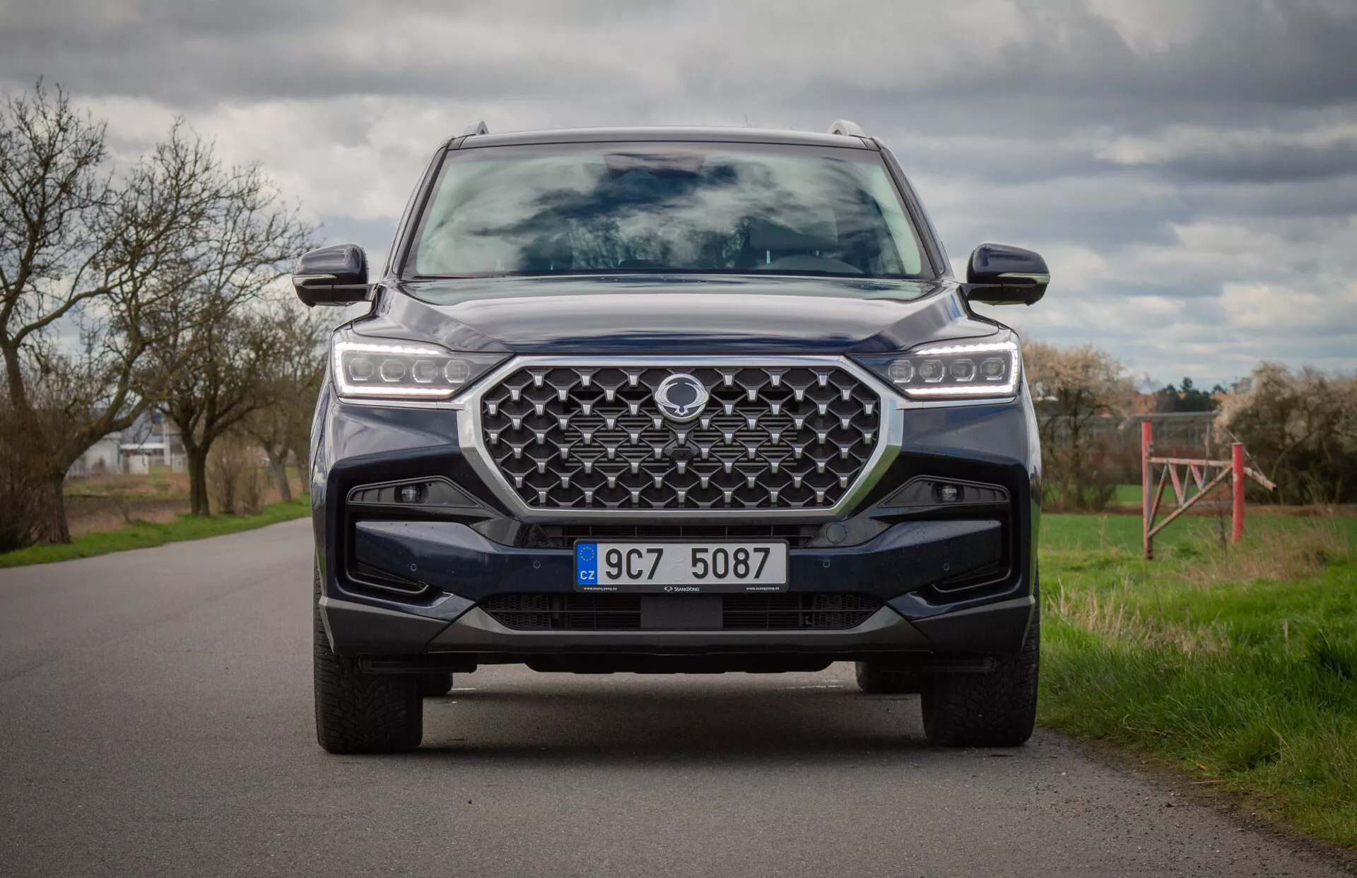 Nowoczesny SUV SsangYong Rexton 2021 w kolorze, uchwycony z pozycji klęczącej ukazującej przednią część pojazdu, reflektory LED, dużą zaprojektowaną atrapę chłodnicy, przedni zderzak z płytą ochronną i zaawansowane systemy asysty kierowcy
