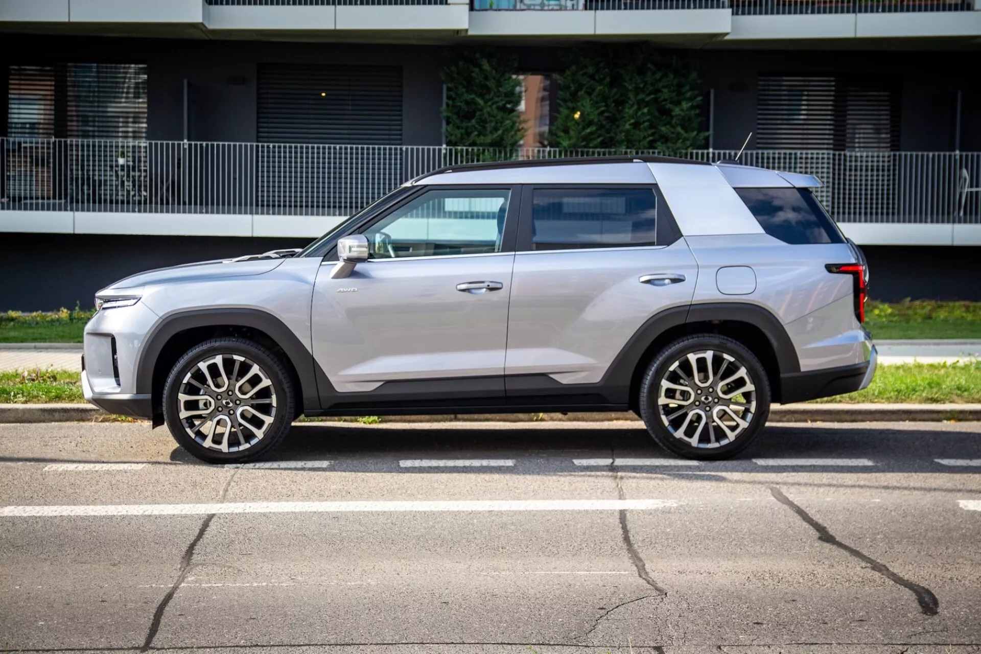 Widok boczny SsangYong Torres 2023 ukazujący jego kanciasty, a jednak aerodynamiczny design, wysoką linię pasa, płaski dach i jasną metaliczną farbę z pięcioramiennymi felgami ze stopu, lusterka boczne w kolorze nadwozia z zintegrowanymi kierunkowskazami, lekko przyciemniane szyby, klamki drzwi wpuszczane i czarne obicie na słonecznej ulicy