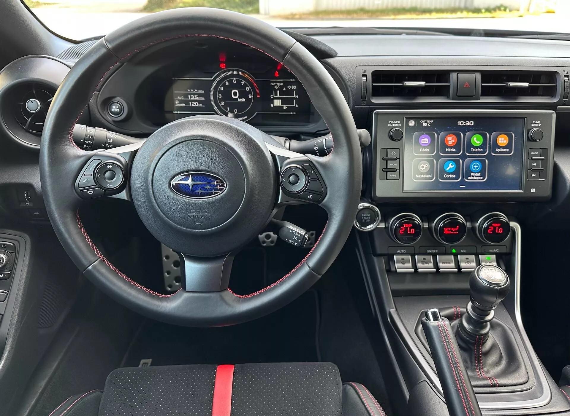 Wnętrze Subaru BRZ 2023 z perspektywy kierowcy pokazujące sportową kierownicę ze skóry z multifunkcyjnymi przyciskami i czerwonymi szwami, zespół wskaźników z analogowymi tarczami i wyświetlaczem cyfrowym, konsolę centralną z dużym ekranem dotykowym systemu rozrywki i kontrolą klimatu dwustrefową, oraz manualną dźwignię zmiany biegów z pokrywą ze skóry i czerwonymi akcentami, na tle czarnego wnętrza z sportowymi czerwonymi akcentami, podkreślające zaawansowane funkcje pojazdu i technologię.