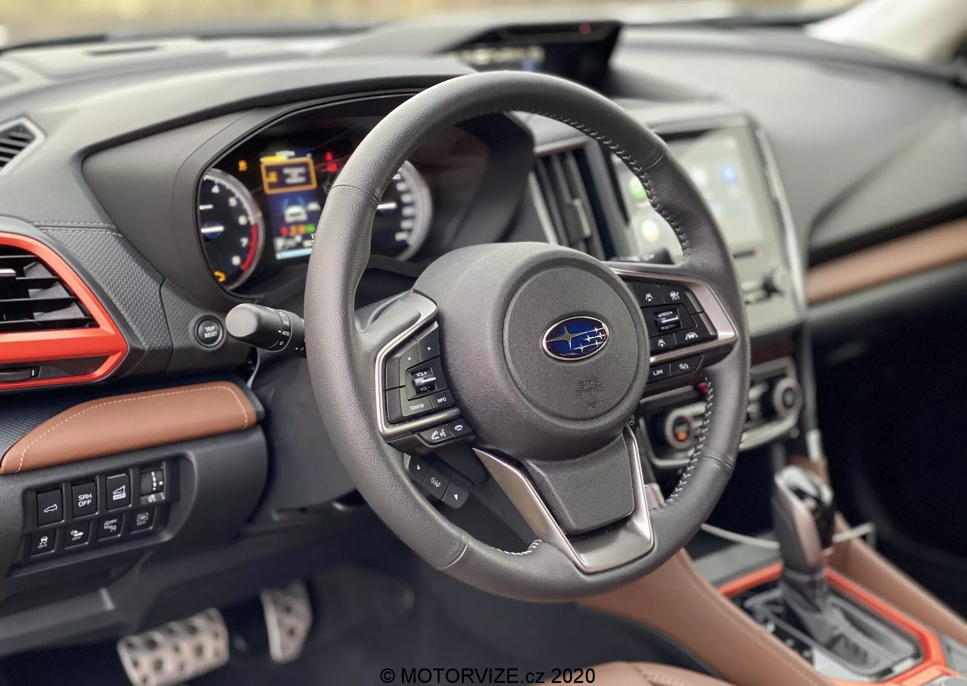 Wnętrze Subaru Forester 2020 z perspektywy kierowcy, pokazujące nowoczesną kierownicę z trzema ramionami z logo Subaru i przyciskami sterującymi, cyfrowy zestaw wskaźników z obrotomierzem, prędkościomierzem i wielofunkcyjnym wyświetlaczem, częściowo widoczną konsolę środkową z systemem informacyjno-rozrywkowym i deską rozdzielczą z obszyciem skórzanym, chromowanymi akcentami i kontrastującymi przeszyciami.