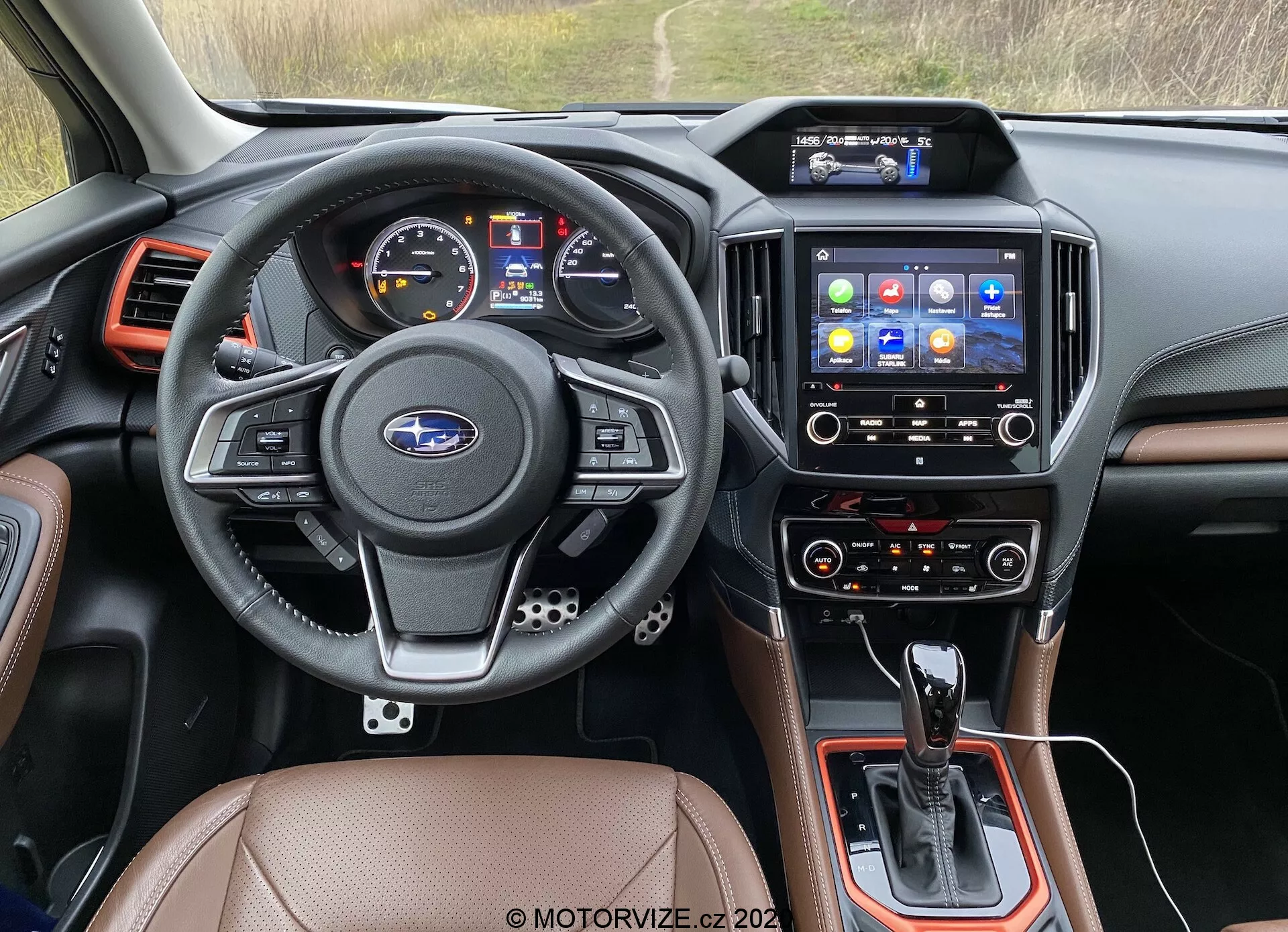 Zdjęcie wnętrza z perspektywy siedzenia kierowcy Subaru Forester 2020, z uwzględnieniem deski rozdzielczej, kierownicy, systemu infotainment i konsoli centralnej. Trójramienna, skórzana kierownica zawiera wielofunkcyjne przyciski do sterowania dźwiękiem, telefonem, komendami głosowymi i tempomatem, z centralnie umieszczonym logo Subaru. Zestaw wskaźników obejmuje cyfrowy wyświetlacz i analogowe mierniki prędkości i obrotów. Konsola centralna mieści dźwignię zmiany biegów automatycznej skrzyni, dodatkowe przyciski i ewentualnie selektor trybu jazdy. Dotykowy ekran infotainment z różnymi ikonami aplikacji oferuje nawigację, multimedia, łączność telefoniczną i ustawienia pojazdu. Poniżej znajdują się przyciski klimatyzacji, sugerujące dwustrefowe ustawienia dla kierowcy i pasażera z przodu. Wykończenia wnętrza obejmują skórzane lub imitujące skórę siedzenia, czarne plastikowe elementy i metaliczne akcenty. Wyloty powietrza otaczają system infotainment i krawędzie deski rozdzielczej. Obraz ten klasyfikuje się jako wnętrza samochodów, technologia motoryzacyjna, deski rozdzielcze, systemy infotainment i sterowanie klimatyzacją.