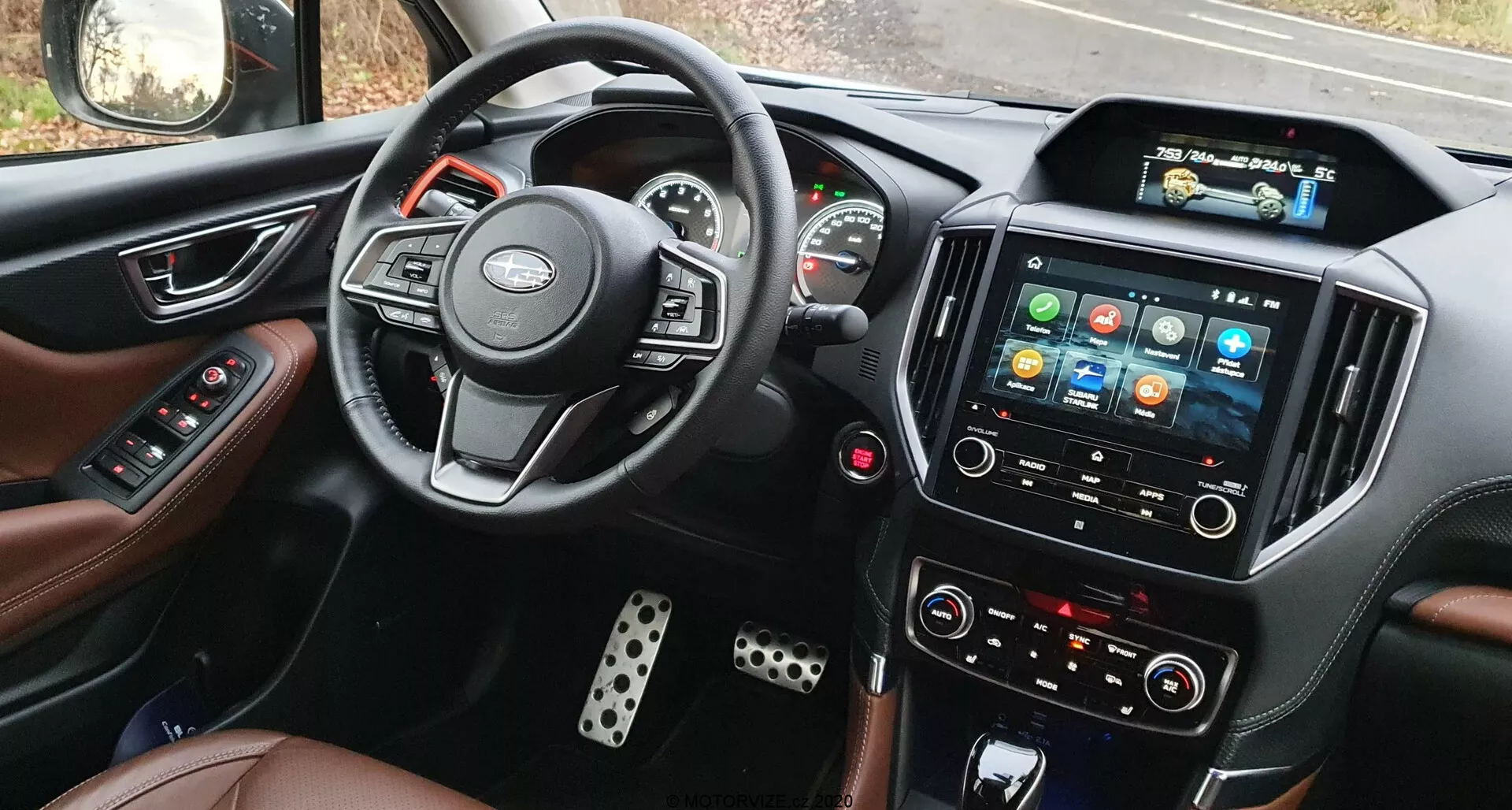 Widok wnętrza Subaru Forester 2020 z miejsca kierowcy, ukazujący kierownicę z zamontowanymi przyciskami, częściowo widoczny zestaw wskaźników z analogowymi miernikami i centralnym wyświetlaczem cyfrowym, wyraźny system infotainment z kolorowymi ikonami na ekranie dotykowym, ręczne przyciski sterowania klimatem, skórzane fotele i metalowe osłony pedałów.