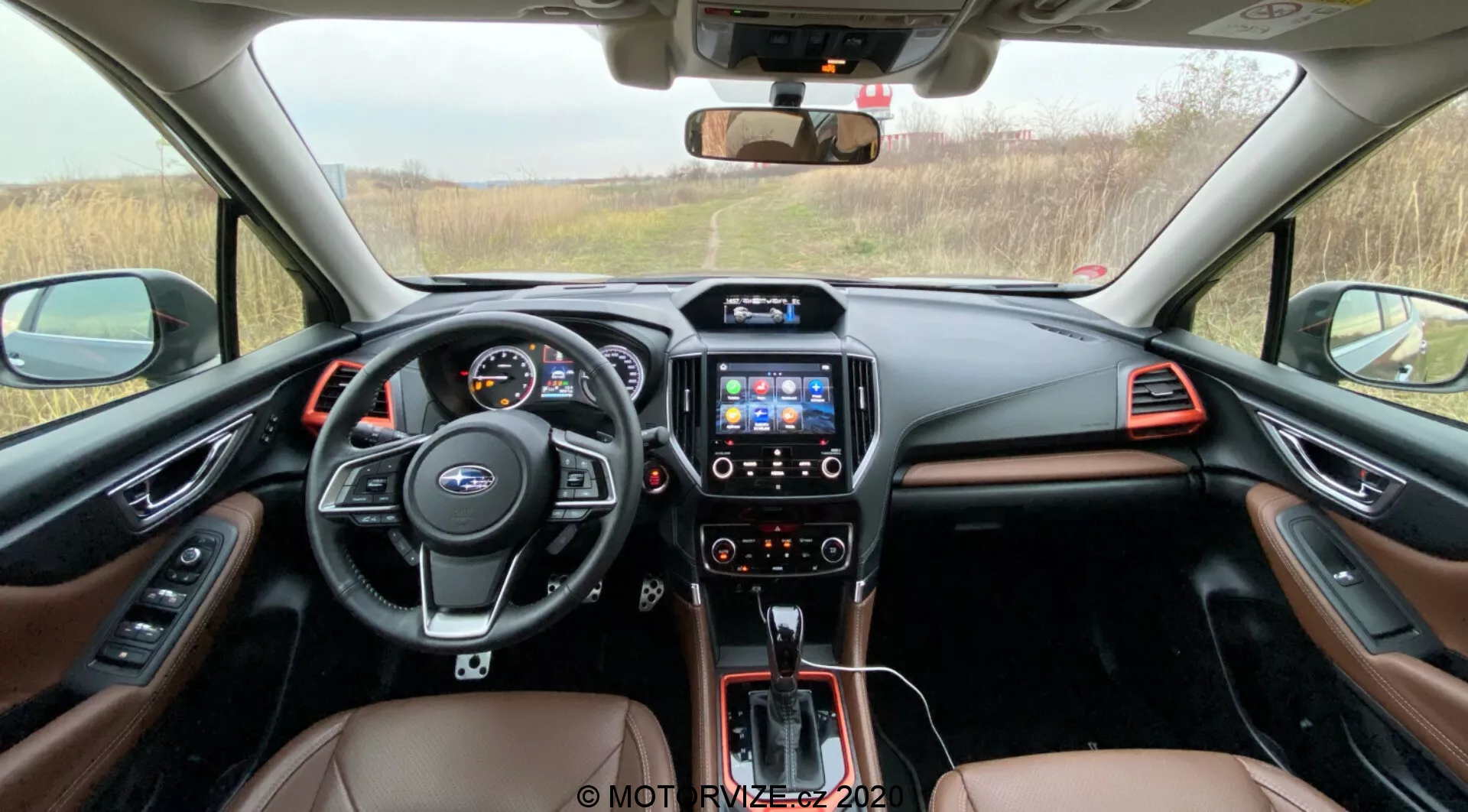 Widok wnętrza przedniej kabiny Subaru Forester 2020, ukazujący wielofunkcyjną kierownicę z przyciskami sterowania, cyfrowy panel instrumentów, konsolę środkową z systemem multimedialnym na ekranie dotykowym, automatyczną dźwignię zmiany biegów i dwukolorowe wnętrze z brązową skórą i metalicznymi wykończeniami, widoczny z tylnych siedzeń w kierunku deski rozdzielczej.
