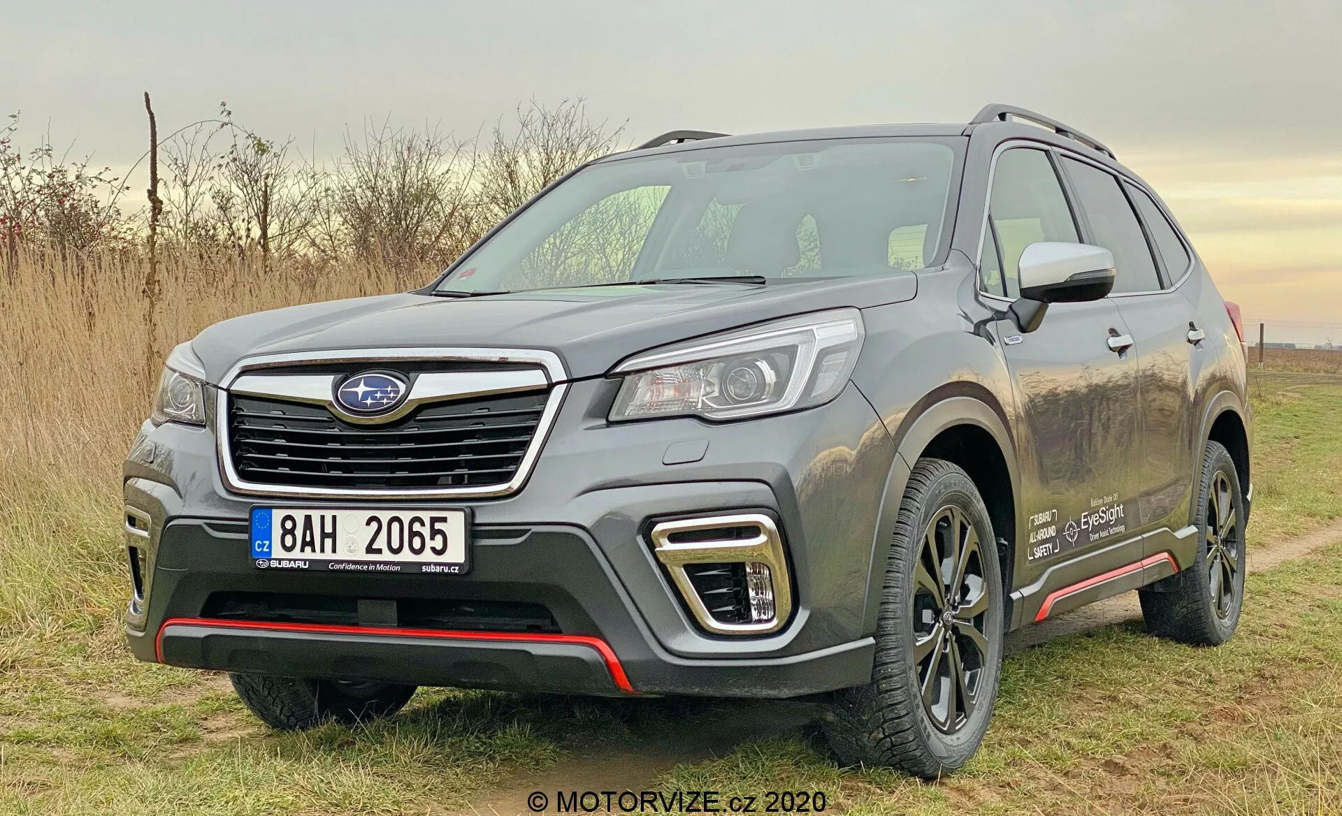 Zdjęcie przedstawia srebrnego SUV Subaru Forester 2020 zaparkowanego na zewnątrz w ciągu dnia w naturalnym świetle. Uchwycone pod kątem trzech czwartych z przodu, zdjęcie wyraźnie pokazuje lewą przednią stronę pojazdu, w tym sześciokątną kratkę z emblematem producenta, eleganckie reflektory LED lub HID, dzienne światła w kształcie litery C wokół świateł przeciwmgielnych oraz atletyczny przedni zderzak z srebrnymi elementami osłonowymi. Rzeźbiona maska posiada podniesione grzbiety, a poszerzone nadkola nadają mu muskularny wygląd. Profil boczny zawiera wyraźną linię charakteru, czarne plastikowe osłony u dołu drzwi, pokryte brudem felgi aluminiowe, relingi dachowe i częściowo widoczne lusterko boczne z zintegrowanymi kierunkowskazami. Na przednim błotniku znajduje się odznaka, która wskazuje na specjalną wersję wyposażenia lub typ silnika. Otoczenie sugeruje lekkie użytkowanie terenowe, podkreślone przez rozpryski błota na bokach pojazdu.