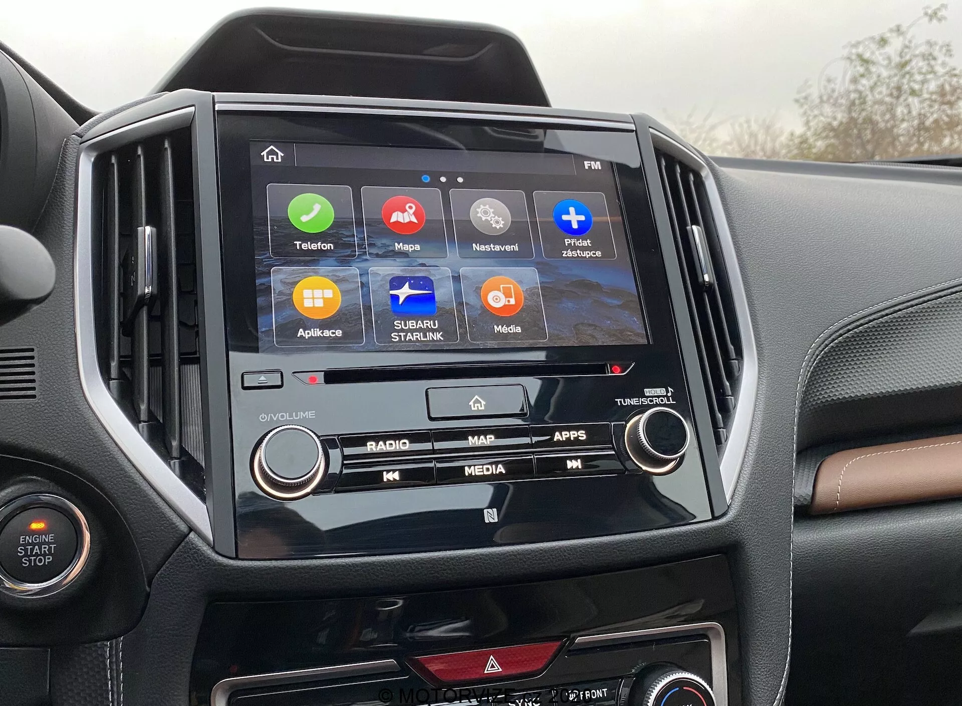 Obraz pokazuje centralną konsolę Subaru Forester 2020, podkreślając system infotainment i wnętrze. W centrum obrazu znajduje się ekran dotykowy z ikonami 'Telefon', 'Mapy', 'Teraz odtwarzane', 'Wiadomości', 'Subaru Starlink', 'Media' i 'Aplikacje', wskazując na różnorodne funkcje takie jak łączność, nawigacja i rozrywka. Ekran infotainment jest osadzony w błyszczącym czarnym obramowaniu, kontrastującym z dwukolorową deską rozdzielczą. Widoczne są również fizyczne przyciski i pokrętła klimatyzacji, ilustrujące kombinację sterowania cyfrowego i manualnego.
