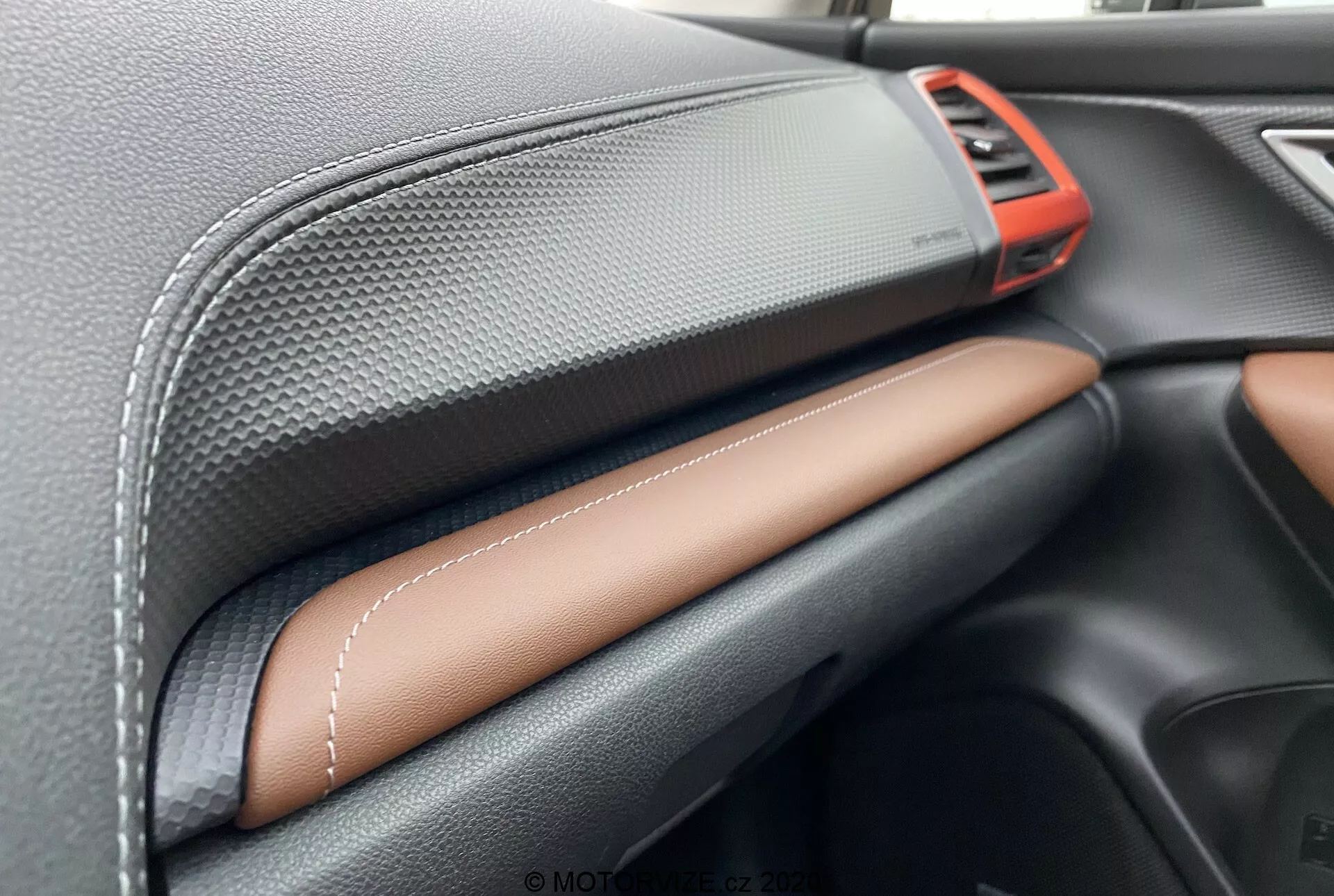 Zbliżenie na wnętrze Subaru Forester 2020 z naciskiem na deskę rozdzielczą. Cechy obejmują jasnoczerwony metalowy nawiew w prawym górnym rogu, czarny materiał o fakturze włókna węglowego na górnej części deski rozdzielczej, kontrastowe szwy na brązowej lub jasnobrązowej skórze na środku oraz elegancki czarny schowek na rękawiczki poniżej. Ukazuje to rzemiosło i jakość materiałów premium.