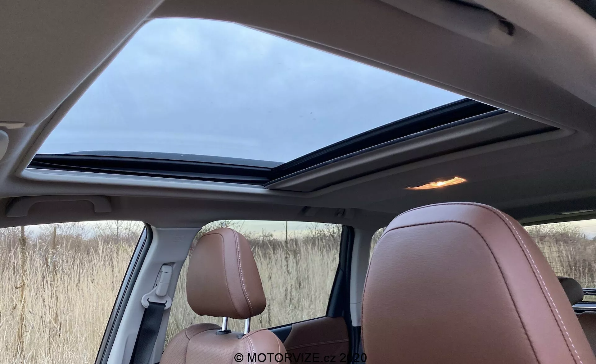 Widok wnętrza Subaru Forester 2020 z perspektywy tylnego pasażera, z widocznym dużym panoramicznym szyberdachem w lekko otwartej pozycji, luksusowymi jasnobrązowymi skórzanymi fotelami z detalicznym przeszyciem oraz kombinacją brązowych, czarnych i beżowych elementów wnętrza. Widoczne są także kratka głośnika przy słupku i przyciemnione boczne okna, oświetlone miękkim dziennym światłem przez zachmurzone niebo.