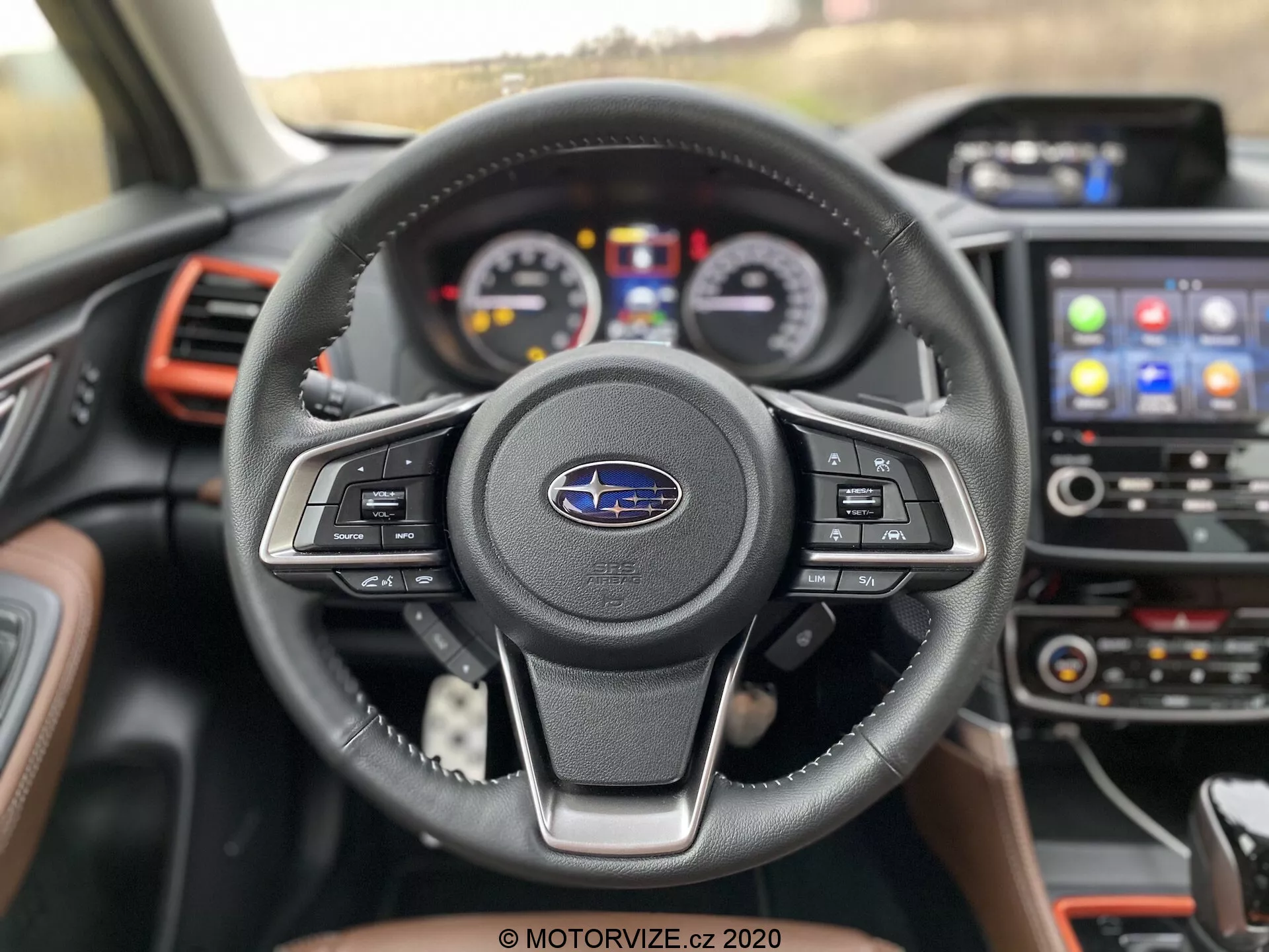 Zbliżony widok wnętrza Subaru Forester 2020 z miejsca kierowcy, z naciskiem na trójramienną, skórzaną kierownicę z wielofunkcyjnymi przyciskami i emblematem Subaru. Za kierownicą widoczny jest zestaw wskaźników z analogowymi tarczami i centralnym wyświetlaczem cyfrowym. Konsola centralna zawiera system infotainment z ekranem dotykowym wyświetlającym ikony nawigacji i łączności telefonicznej. Czerwone akcenty na otworach wentylacyjnych i desce rozdzielczej dodają sportowego charakteru nowoczesnemu, dobrze wyposażonemu pojazdowi.