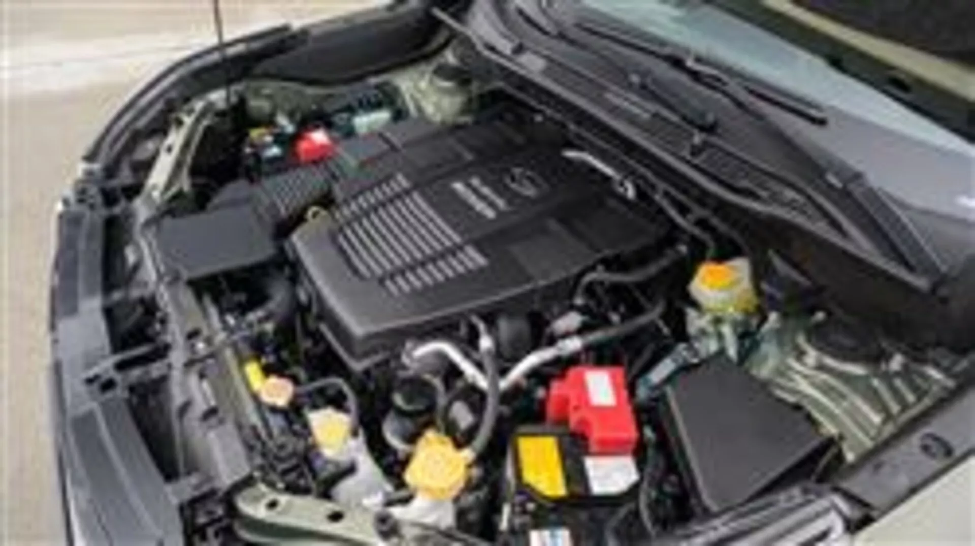 Zbliżenie na komorę silnika Subaru Forester z 2015 roku z przodu od strony kierowcy, pokazujące pokrywę silnika z logo, pojemnik na płyn chłodzący z żółtą nakrętką i czerwone pokrycie dodatniego bieguna baterii wśród innych komponentów mechanicznych i elektrycznych