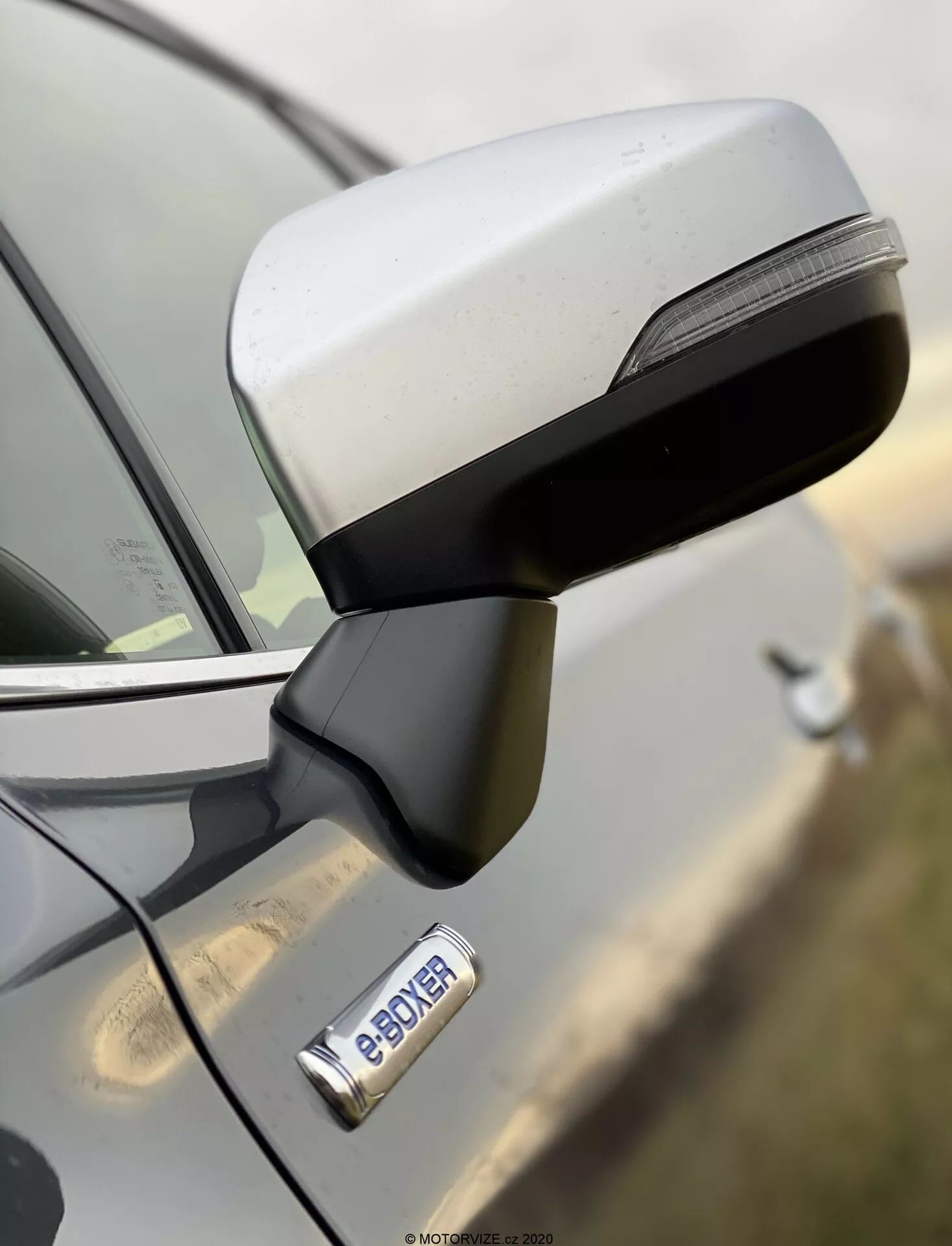 Zbliżenie na lusterko boczne i przednie drzwi Subaru Forester 2020 z widoku bocznego, podkreślające aerodynamiczny kształt lusterka z wbudowanymi kierunkowskazami LED oraz chromowaną klamkę drzwi z przyciskiem do bezkluczykowego dostępu. Odznaka 'e-POWER' wskazuje na napęd hybrydowy lub elektryczny pojazdu. Obraz o wysokiej rozdzielczości z małą głębią ostrości, podkreślający lusterko i odznakę, podczas gdy tło jest rozmazane.