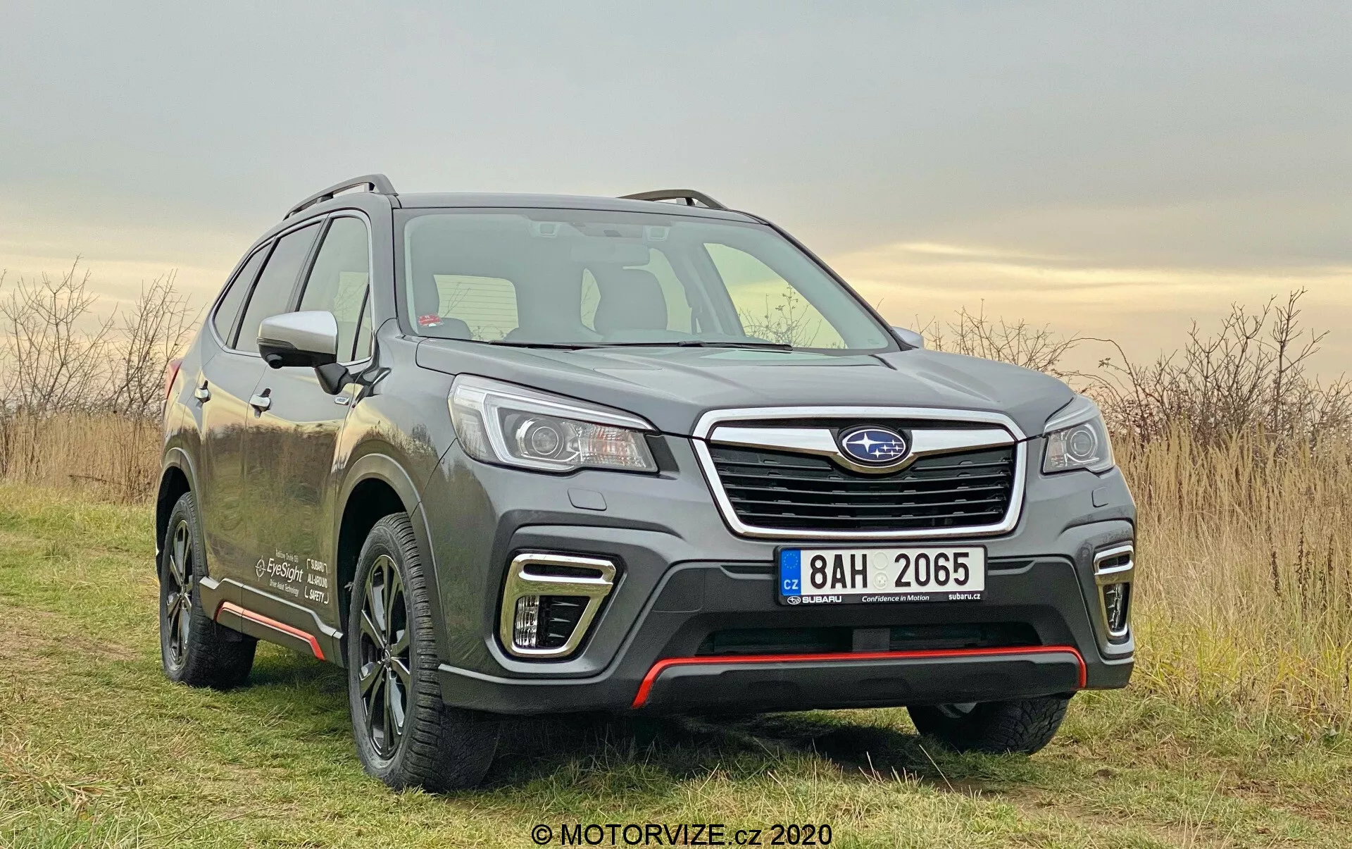 Subaru Forester 2020 na zdjęciu, nowoczesne SUV uchwycone pod kątem trzech czwartych z przodu. Umieszczony nieco na prawo od ramy na nieutwardzonej drodze z trawą, pod mglistym niebem. Zdjęcie podkreśla kluczowe cechy takie jak przednia fasada, krata z chromowaną poziomą listwą i modułem czujników, szczegółowe reflektory LED, konturowane linie maski i profil boczny kierowcy. Widoczna felga aluminiowa z wieloramiennym wzorem, lusterka boczne z zintegrowanymi kierunkowskazami i linie nadwozia wzdłuż drzwi sugerują wyższy poziom wykończenia. Błoto na pojeździe sugeruje jego użycie w terenie i na nieutwardzonych drogach. Miękkie światło dzienne i pochmurna pogoda zapewniają równomierne oświetlenie, podkreślając design i możliwości samochodu.