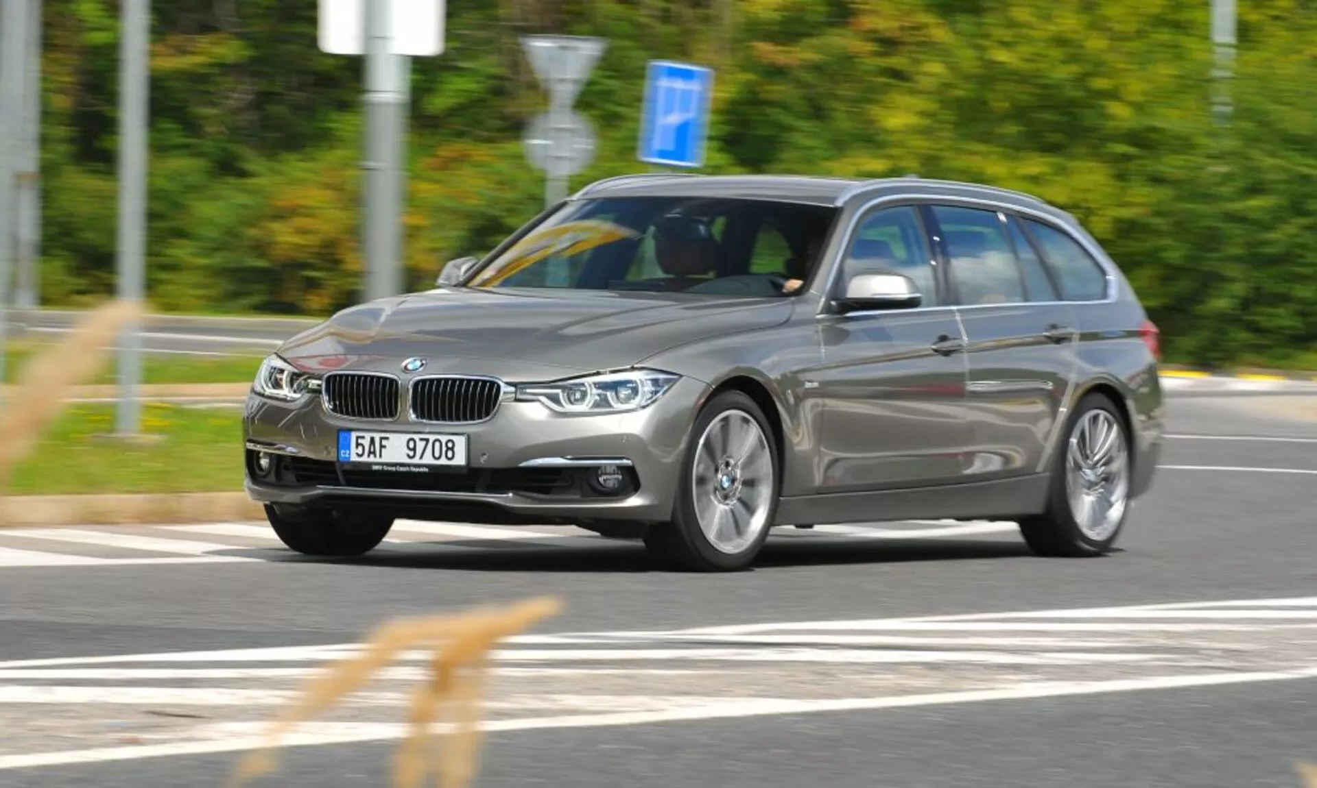 Dynamiczny trzykwartalny widok z przodu w ruchu srebrnego kombi BMW z charakterystycznym grillem w kształcie nerek i emblematem, uchwycony z lekko podniesionego punktu widzenia, wskazujący na prędkość i ruch, skupiający się na cechach projektowych i technologicznych takich jak kątowe reflektory, felgi aluminiowe i relingi dachowe na zewnętrznym tle z rozmyciem ruchu.