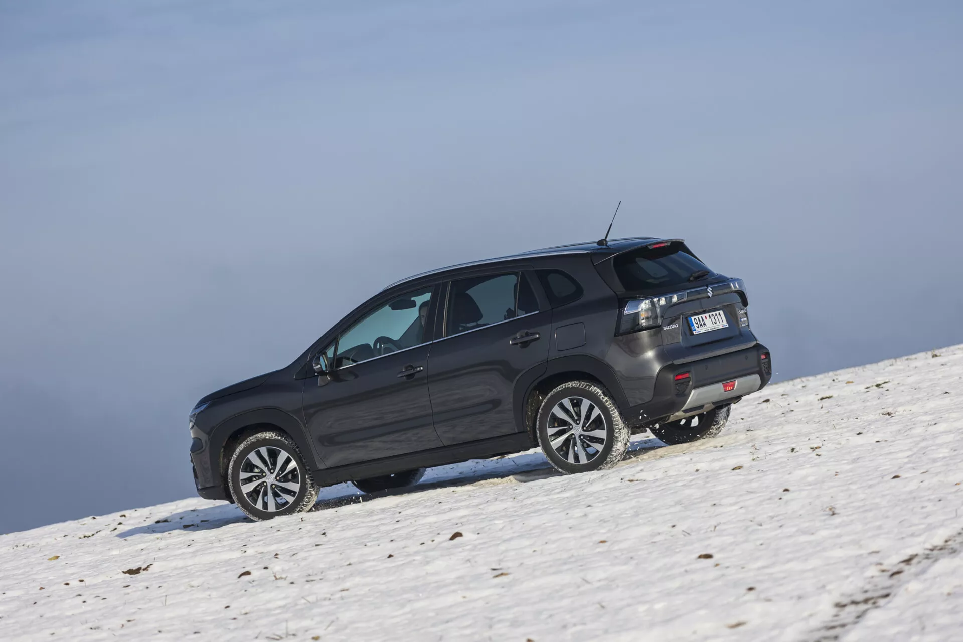 SUV Suzuki S-Cross 2022 w ciemnym odcieniu umieszczony na śnieżnym zboczu, uchwycony z ukośnego tylnego kąta podkreślającego jego boczny i tylny profil, duże koła ze stopów, przyciemniane okna z ogrzewaniem tylnej szyby, światła tylne LED, spojler dachowy, i potencjalne zdolności terenowe w warunkach jasnego nieba.