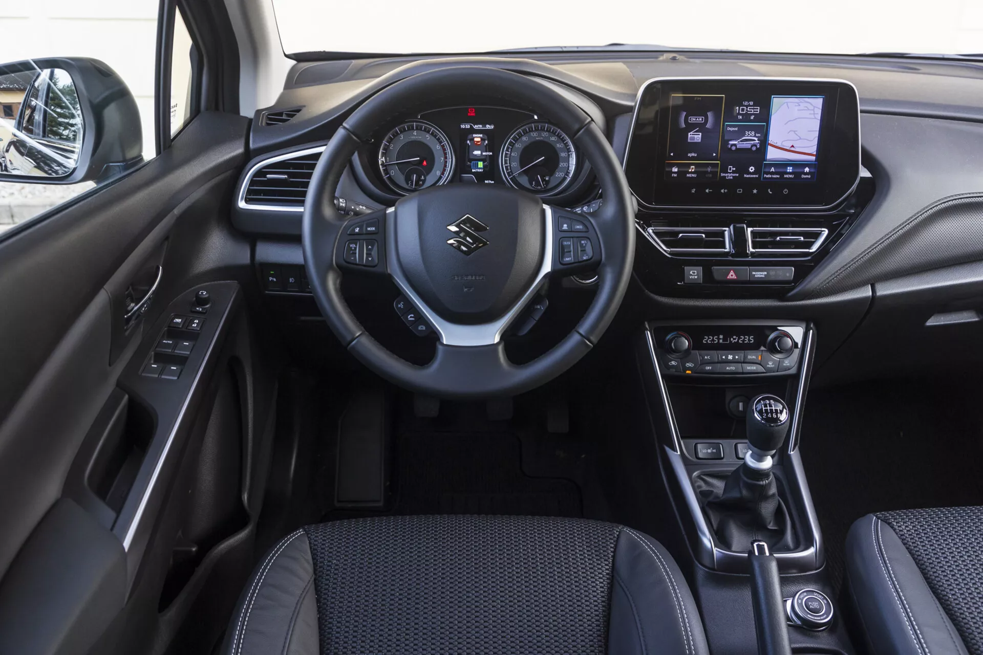 Wnętrze przedniej kabiny Suzuki S-Cross 2022 pokazujące deskę rozdzielczą, kierownicę z zamontowanymi sterowaniami, kombinacje cyfrowo-analogowego zestawu wskaźników, ekran dotykowy systemu infotainment, manualne sterowanie klimatyzacją, skórzaną dźwignię zmiany biegów, selektor trybu jazdy, elektroniczny hamulec postojowy oraz panel drzwiowy z przyciskami sterowania
