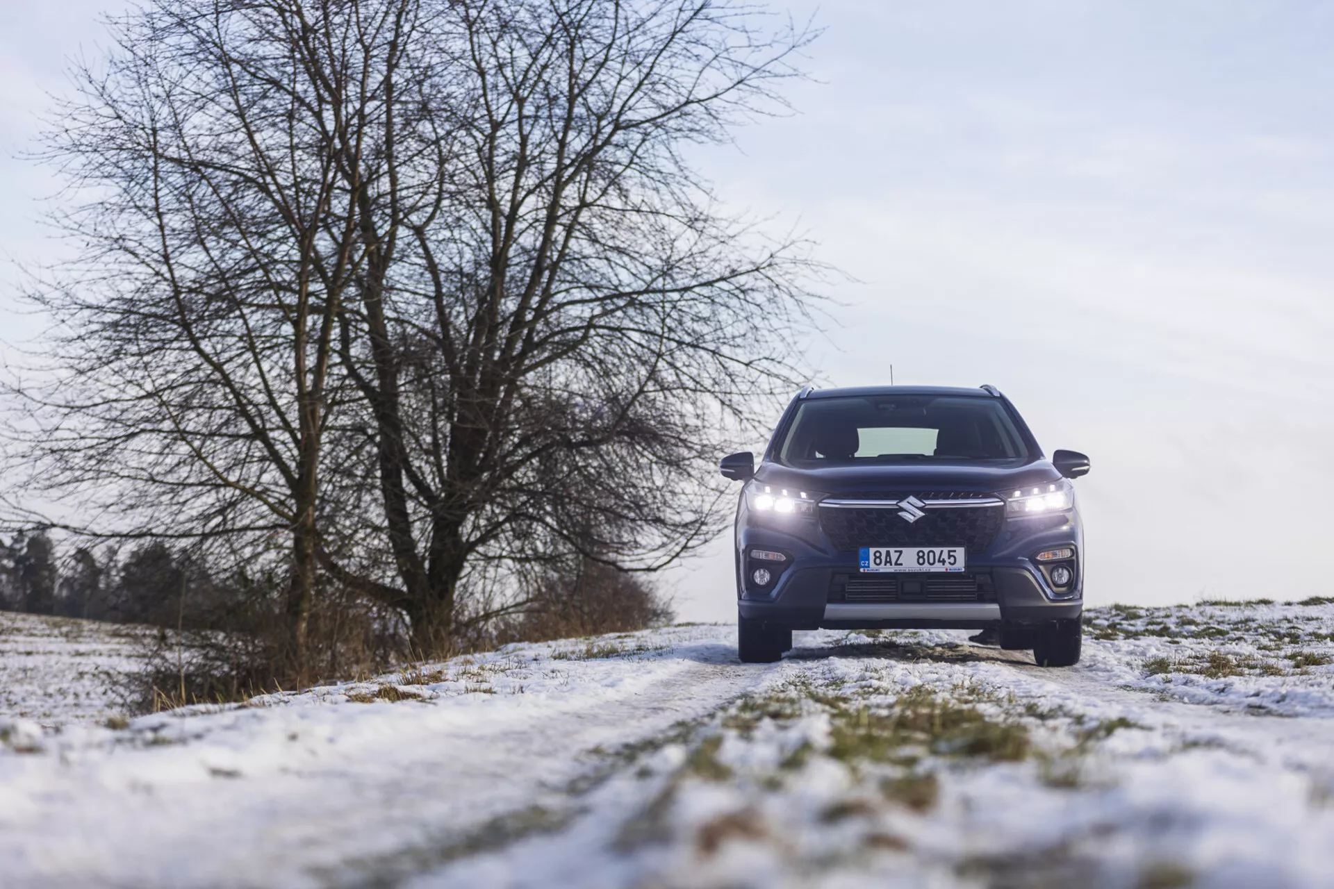 Przedni widok Suzuki S-Cross 2022 na zaśnieżonej wiejskiej drodze, prezentujący reflektory LED, nowoczesny wzór grilla, felgi aluminiowe i elegancką karoserię SUV na tle zimowego krajobrazu z drzewami bez liści i pochmurnym niebem.