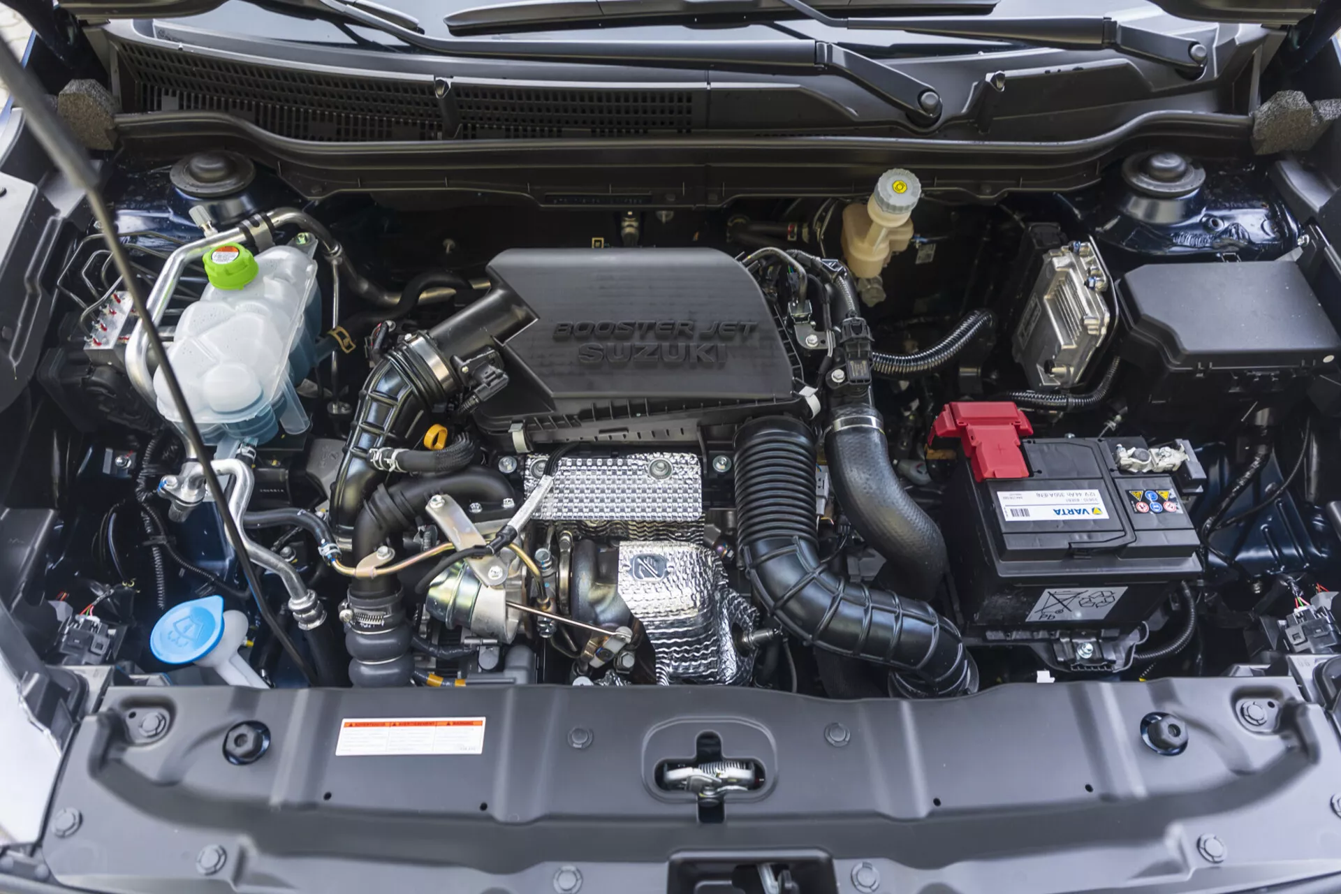Komora silnika Suzuki S-Cross 2022 widziana z przodu z widocznymi elementami takimi jak blok silnika, głowica cylindrów, kolektor dolotowy, oraz cechy technologiczne w tym turbodoładowanie i zmienny czas otwarcia zaworów, razem z brandingiem Suzuki na pokrywie silnika, w nienagannej kondycji.