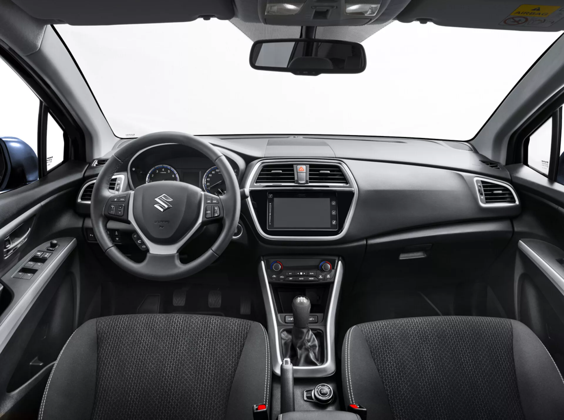 Widok wnętrza Suzuki S-Cross 2022 z tylnego siedzenia przedstawiający deskę rozdzielczą, kierownicę z przyciskami, cyfrowy zestaw wskaźników, system infotainment, sterowanie klimatyzacją, dźwignię automatycznej skrzyni biegów i szczegółowo wykonane przednie fotele z tkaniną
