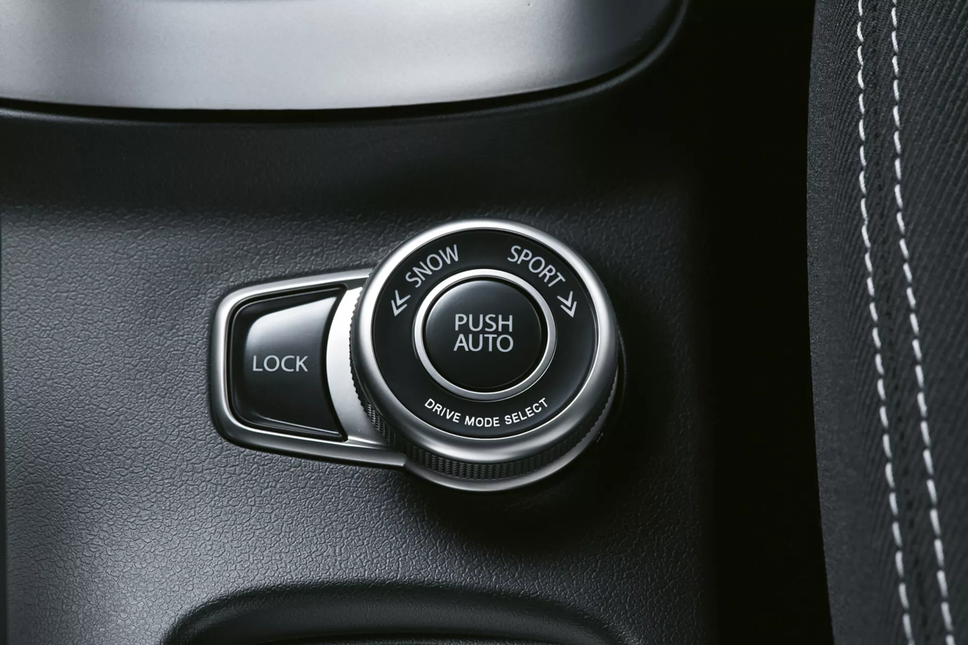 Zbliżenie wnętrza Suzuki S-Cross 2022 pokazujące przełącznik trybu jazdy oznaczony jako 'PUSH AUTO' z opcjami 'SNOW' i 'SPORT', obok przycisku 'LOCK', na panelu wysokiej jakości o ciemnych odcieniach, możliwe z teksturą skóry i widocznymi szwami.