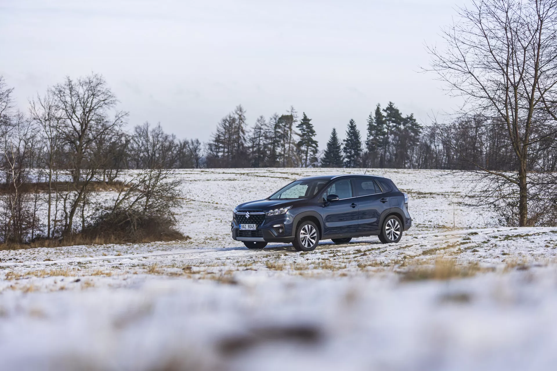 Suzuki S-Cross 2022 w zimowym krajobrazie ze śniegiem, prezentujące jego dynamiczny design, zaawansowane oświetlenie, felgi aluminiowe i relingi dachowe, podkreślające styl SUV i zdolności terenowe.