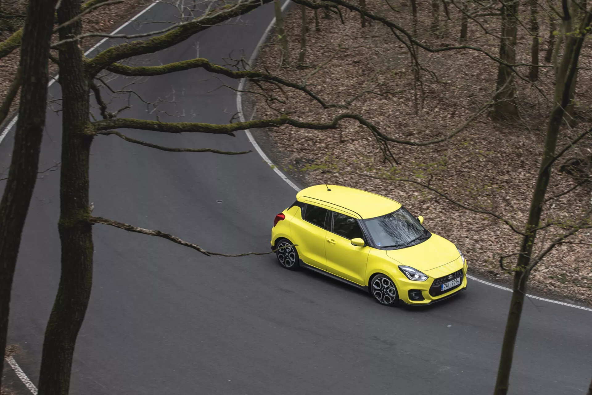 Widok z lotu ptaka na jaskrawożółty Suzuki Swift 1995 z dwukolorową kolorystyką, podróżujący krętą drogą przez las, prezentujący felgi aluminiowe, nowoczesne reflektory LED i kompaktową konstrukcję typu hatchback.