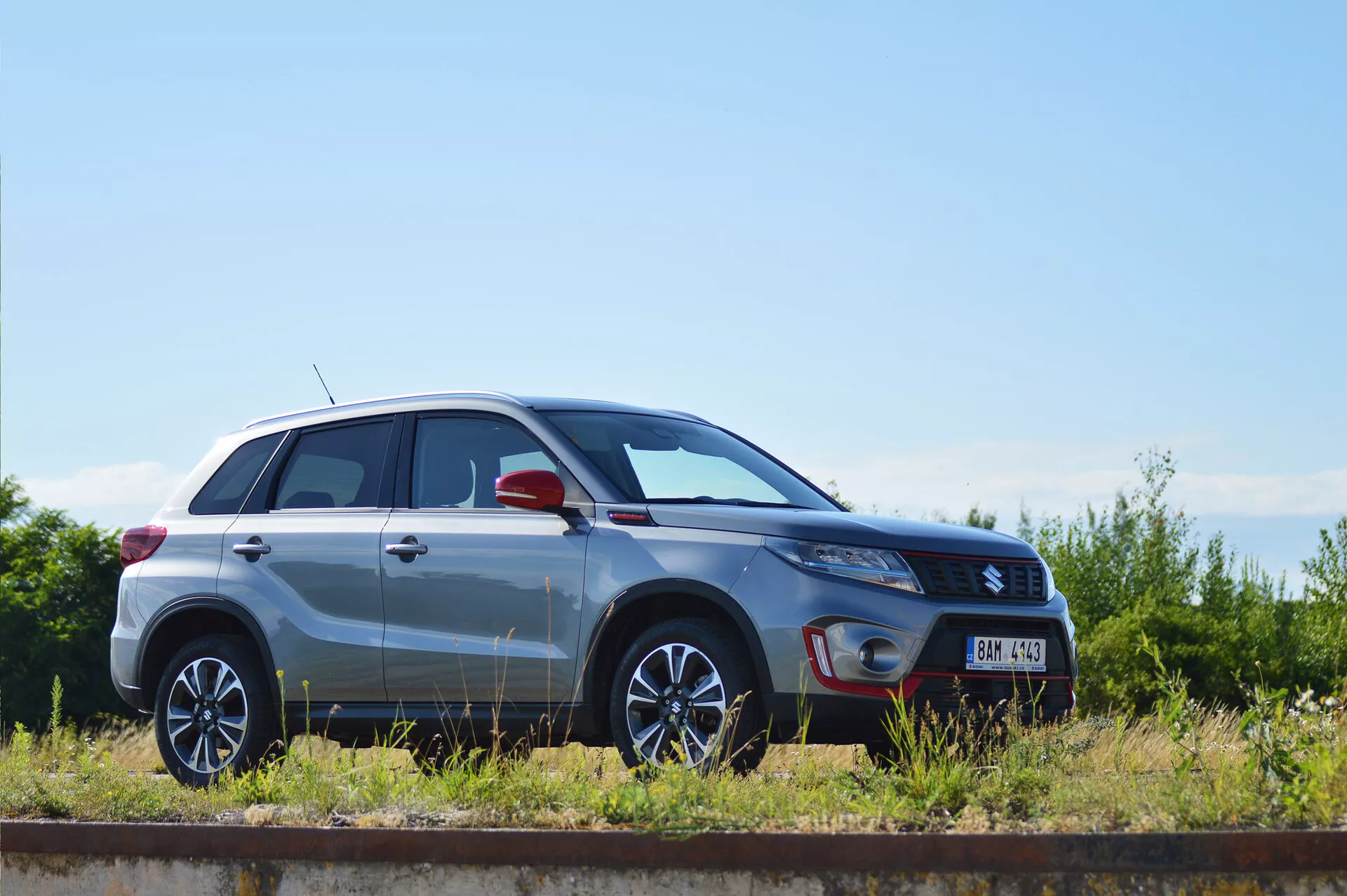 Widok z profilu bocznego srebrnego kompaktowego SUV-a Suzuki Vitara z 2018 roku, zaparkowanego równolegle do torów kolejowych w słoneczny dzień. Wyraźnie widoczne są linie projektowe, aluminiowe felgi z wieloramiennym wzorem, metaliczne wykończenie lakieru, zamknięte drzwi i okna, kształt reflektorów, lusterko boczne z potencjalnie zintegrowanymi kierunkowskazami oraz plakietka poziomu wykończenia, co ukazuje design i cechy technologiczne pojazdu.