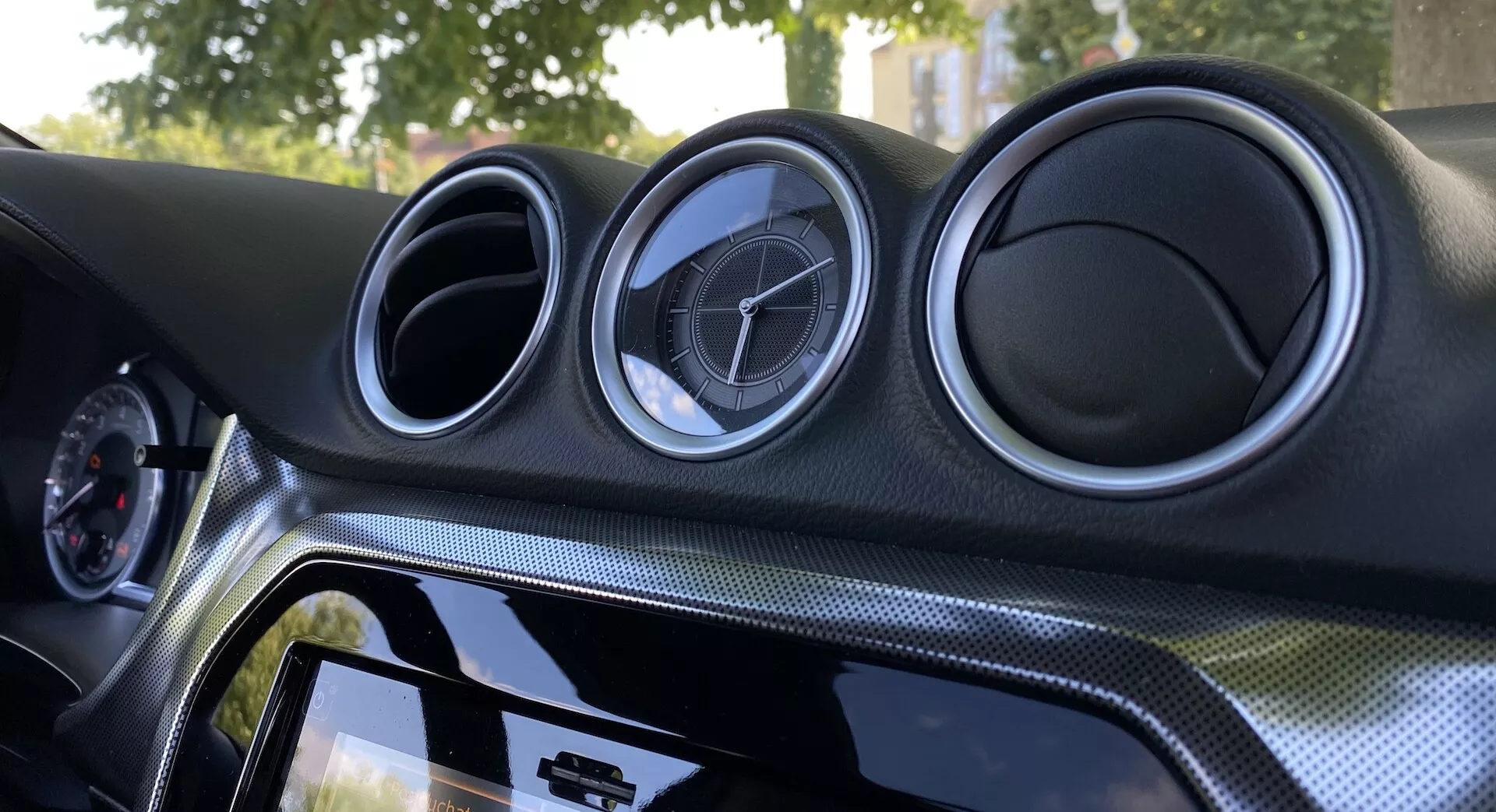Zbliżenie wnętrza Suzuki Vitara 2018 z naciskiem na centralny panel deski rozdzielczej. Deska rozdzielcza zawiera trzy okrągłe metalicznie wyglądające otwory wentylacyjne z emblematem producenta, dotykowy system informacyjno-rozrywkowy z błyszczącą czarną ramką, materiał imitujący włókno węglowe oraz oświetlenie ambientowe dla lepszego wyglądu.