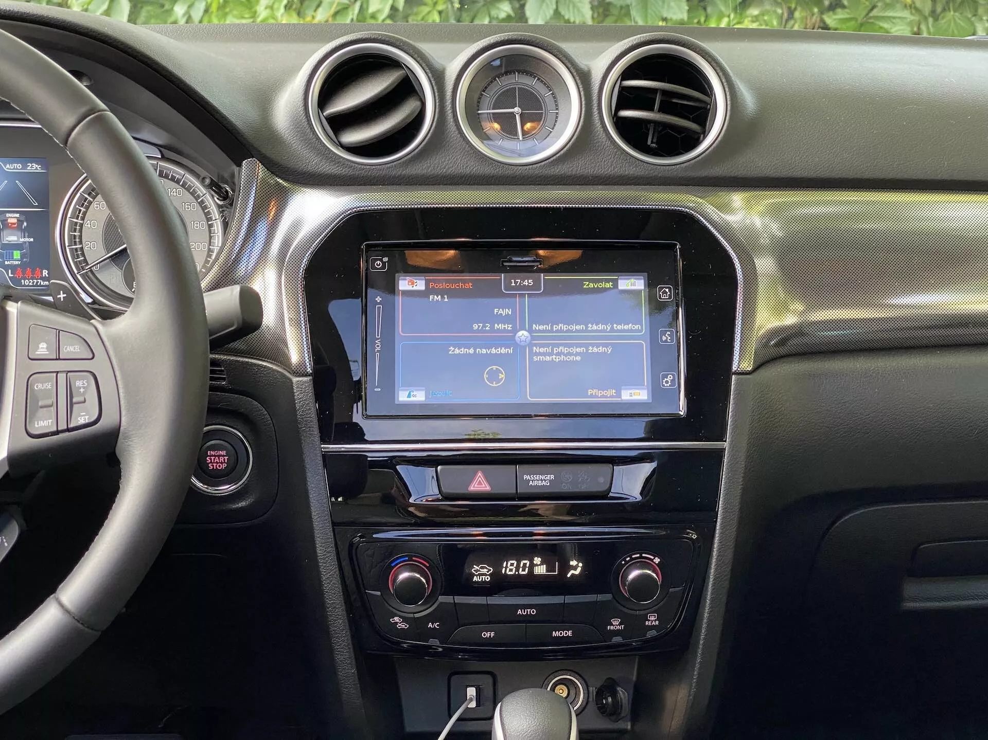 Widok wnętrza Suzuki Vitara 2018 koncentrujący się na centralnej desce rozdzielczej. Sekcja zawiera częściowo widoczną kierownicę po lewej stronie z wieloma przyciskami kontrolnymi, prawdopodobnie do sterowania audio, połączeń bez użycia rąk i tempomatu. Centralna konsola wyświetla ekran dotykowy systemu infotainment, prawdopodobnie pokazujący nawigację lub interfejs menu, otoczony pionowymi otworami wentylacyjnymi i zwieńczony trzema okrągłymi otworami. Poniżej ekranu dotykowego znajduje się rząd przycisków i pokręteł umożliwiających szybki dostęp do ustawień infotainmentu i pojazdu. Regulatory klimatyzacji są umieszczone niżej, co sugeruje dwustrefowy system klimatyzacji. Deska rozdzielcza posiada miękki materiał z widocznymi szwami, a okrągły zegar analogowy jest zamontowany między centralnymi okrągłymi otworami wentylacyjnymi. Widoczność jest skoncentrowana na funkcjach deski rozdzielczej, bez okien lub przedniej szyby w zasięgu wzroku.