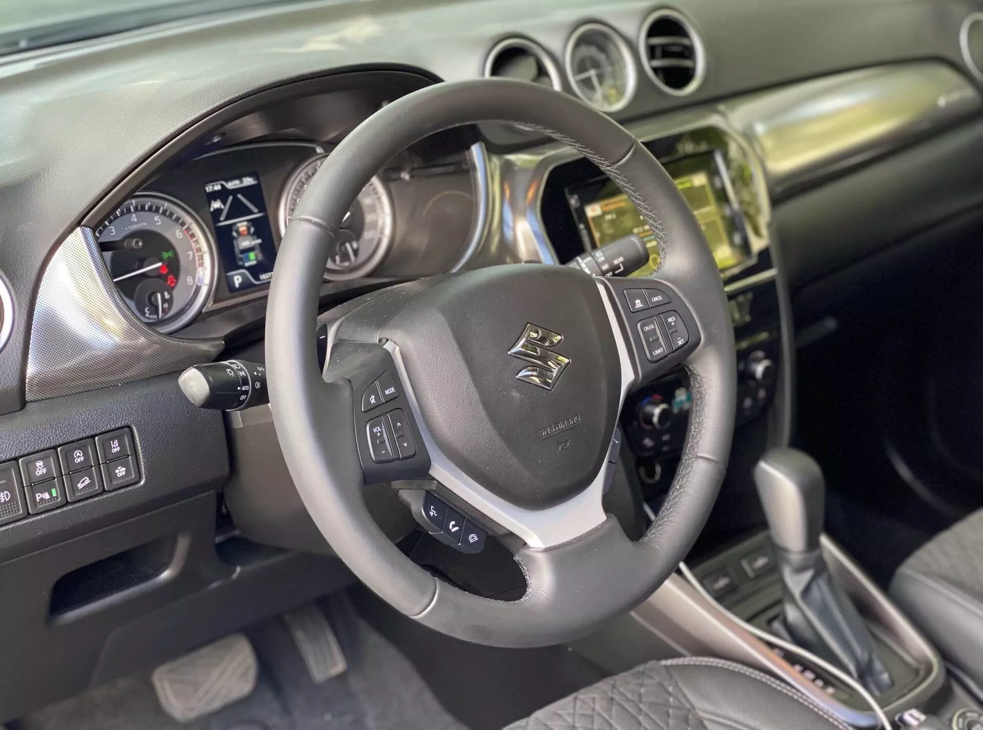 Wnętrze Suzuki Vitara 2018, uchwycenie perspektywy kierowcy. Zbliżenie na wielofunkcyjną kierownicę pokrytą skórą z emblematem na środku, z zamontowanymi sterownikami dźwięku, sterowania głosem i tempomatu z metalicznymi akcentami. Za nią zestaw wskaźników z analogowymi tarczami do prędkościomierza i obrotomierza oraz ekranem cyfrowym do informacji o pojeździe, sugerujący, że samochód jest wyłączony lub w trybie akcesoryjnym. Konsola centralna pokazuje stos wykończony metalem lub chromem z systemem infotainment z ekranem dotykowym, przyciskami i pokrętłami do sterowania klimatem i pojazdem. Automatyczna dźwignia zmiany biegów z elementami skórzanymi sugeruje różne tryby jazdy. Wnętrze łączy skórę i szczotkowany metal, odzwierciedlając nowoczesny design. Domyślnie zaawansowane funkcje obejmują Bluetooth, nawigację, asystenty parkowania i systemy wspomagania kierowcy.