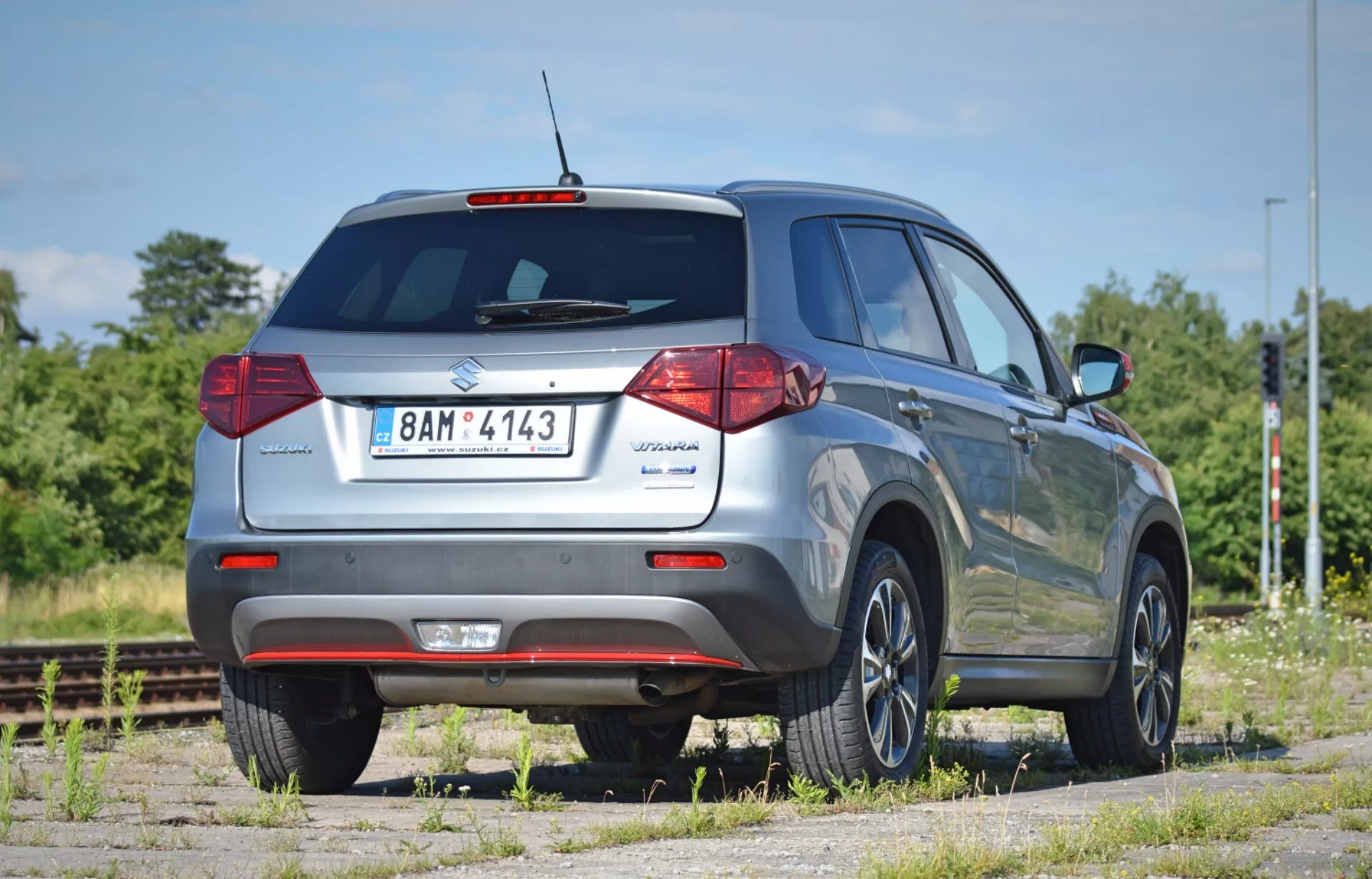 Widok tylni SUV-a Suzuki Vitara 2018 z niskiego kąta, podkreślający jego wysokość i prześwit. Pojazd ustawiony jest pod kątem 45 stopni, zdjęcie ukazuje tylne drzwi z oznaczeniem modelu, tylne szyby z wycieraczką, tradycyjne halogenowe światła tylne, odblaskowy tylny zderzak oraz widoczna rura wydechowa. Widoczne elements prawego boku obejmują tylne drzwi pasażera, boczne okna, relingi dachowe i standardowe felgi aluminiowe. Brak widocznych kierunkowskazów zintegrowanych z lusterkami bocznymi. Pojazd zaparkowany jest na kurzystej powierzchni obok torów kolejowych, w suchej, słonecznej okolicy z zielenią i bezchmurnym niebem.