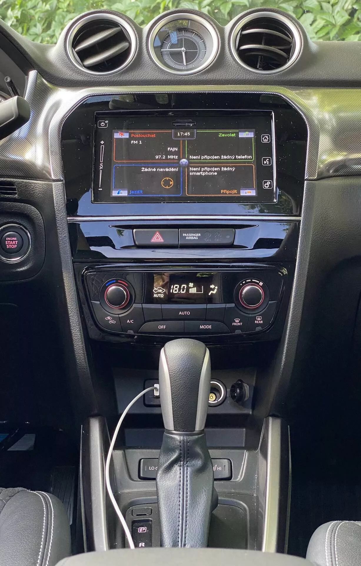 Szczegółowy widok wnętrza Suzuki Vitara 2018, podkreślający konsolę środkową i deskę rozdzielczą z perspektywy przednich siedzeń. Obraz podkreśla dotykowy ekran systemu multimedialnego wyświetlający menu, system klimatyzacji dwustrefowej z cyfrowymi odczytami i dźwignię automatycznej skrzyni biegów. Wnętrze prezentuje wykończenia w ciemnych kolorach z mieszanką matowych i błyszczących tekstur, ilustrując jakość materiałów i nowoczesną technologię motoryzacyjną.