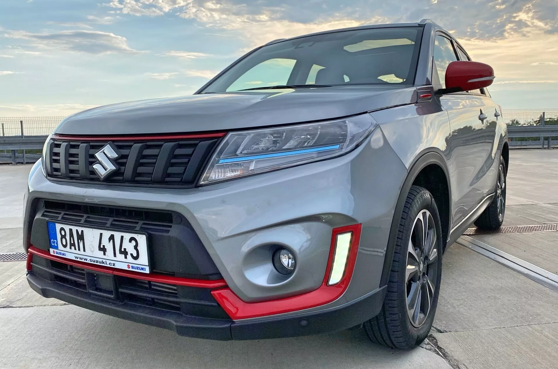 Zbliżony widok boczny nowoczesnego kompaktowego SUV-a, ustawiony pod kątem, aby pokazać część bocznego profilu. Obraz podkreśla solidność pojazdu z widocznymi szczegółami, takimi jak przedni zderzak, grill z poziomymi prętami i emblematem, eleganckie reflektory LED, światła przeciwmgielne z czerwonymi wstawkami, aluminiowe felgi i muskularne błotniki. Ciemne aluminiowe felgi i kontrastowy element wykończeniowy wskazują na wersję premium lub sportową.