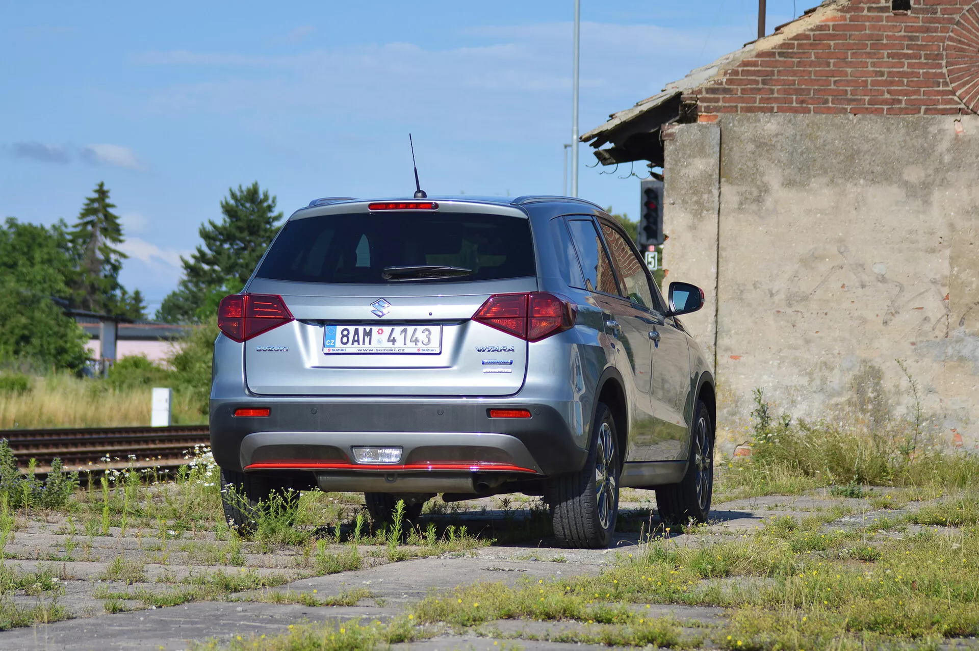 Metaliczny szary SUV Škoda Yeti zaparkowany na zaniedbanym terenie z plamami trawy i betonu, sfotografowany z tylnego trójćwierciowego widoku. Obraz przedstawia charakterystyczny tylni design pojazdu z konwencjonalnymi tylnymi światłami, tylną wycieraczką, wysoko zamontowanym światłem stopu i częścią tylnego spojlera. Logo Škoda jest wyśrodkowane na tylnej klapie, model 'Yeti' po lewej stronie i odznaka '4x4' po prawej stronie, co wskazuje na napęd na cztery koła. Samochód ma pięcioramienne felgi aluminiowe, wydaje się być w dobrym stanie i znajduje się w przemysłowej okolicy z zaniechanymi torami kolejowymi oraz zniszczoną strukturą w tle, w naturalnym świetle słonecznym.