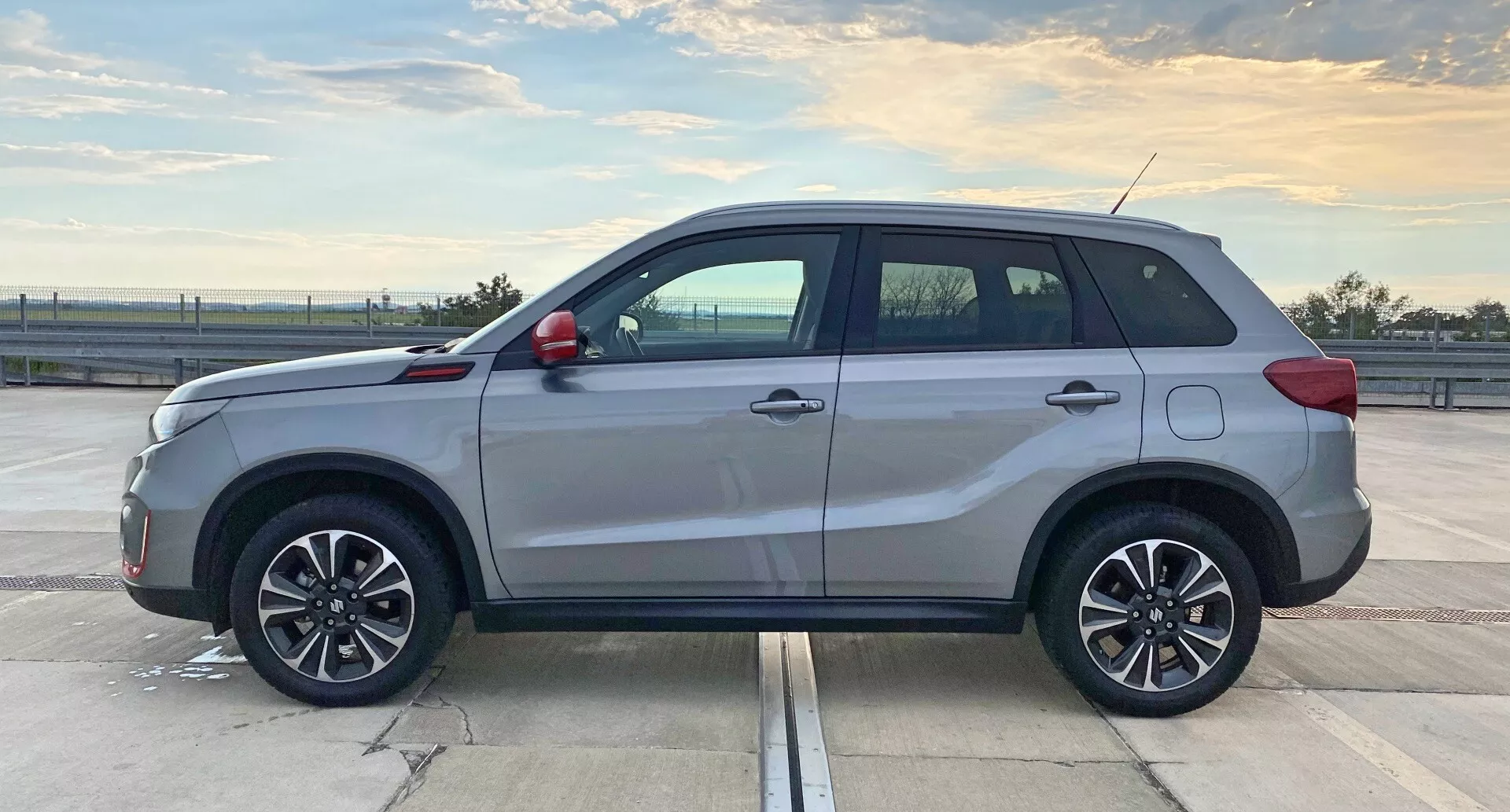 Profil boczny nowoczesnego SUV-a Suzuki Vitara 2018, uchwycony w ciągu dnia na przestronnym parkingu. Skupia się na środkowej części pojazdu, pokazując długość i boczne elementy designerskie takie jak kształty drzwi, progi boczne oraz nadkola. Naturalne oświetlenie uwydatnia pięcioro drzwi, wieloszprychowe felgi aluminiowe, relingi dachowe, kontrastujące klamki i elementy wykończeniowe, antenę oraz potencjalnie przyciemnione szyby. SUV prezentuje się w nienagannym stanie z refleksyjnym połyskiem na karoserii.