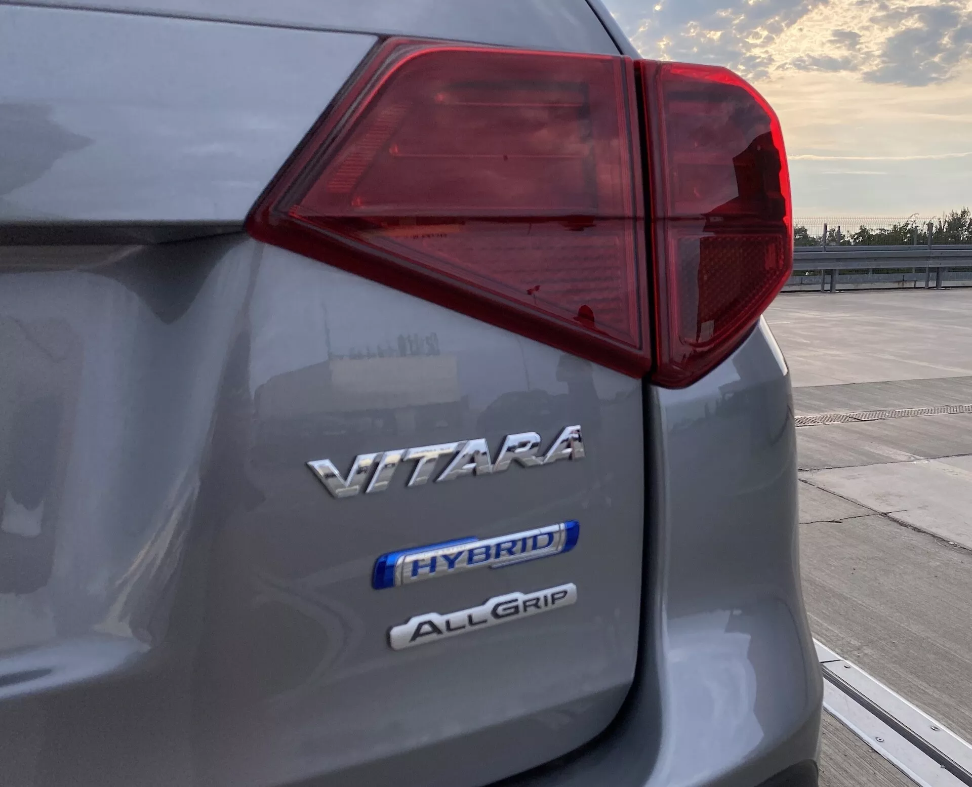 Zbliżenie na lewy tylny róg Suzuki Vitara 2018 ukazujące skomplikowany wzór lampy tylnej LED i trzy oznaczenia wskazujące nazwę modelu 'VITARA', technologię hybrydową 'HYBRID' oraz system napędu na cztery koła 'ALLGRIP', uchwycone o świcie lub zmierzchu na otwartym parkingu.