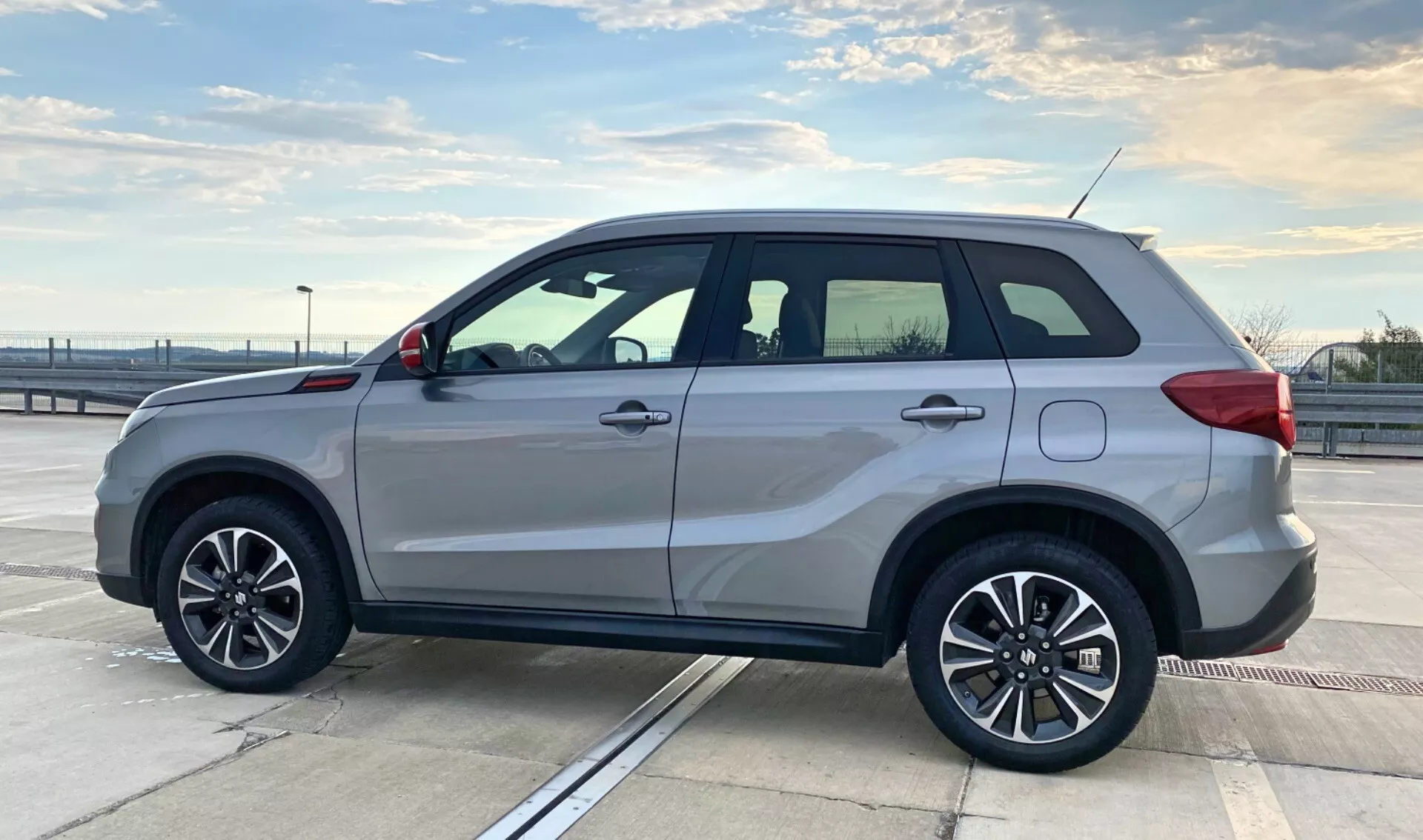 Widok z profilu bocznego na Suzuki Vitara 2018, zaparkowany na górnym poziomie parkingu pod czystym niebem. SUV posiada pięcioramienne aluminiowe koła, srebrne lakierowanie, zintegrowane kierunkowskazy na lusterkach bocznych, relingi dachowe, klamki drzwi w kolorze nadwozia, subtelny boczny stopień oraz przyciemnione okna z chromowanymi ramkami. Design pojazdu podkreśla sportową elegancję i funkcjonalność, z kanciastą karoserią dla maksymalizacji przestrzeni wewnętrznej. Otoczenie plenerowe i kompozycja uwydatniają styl i budowę samochodu, odpowiednie do kategorii związanych z pojazdami.