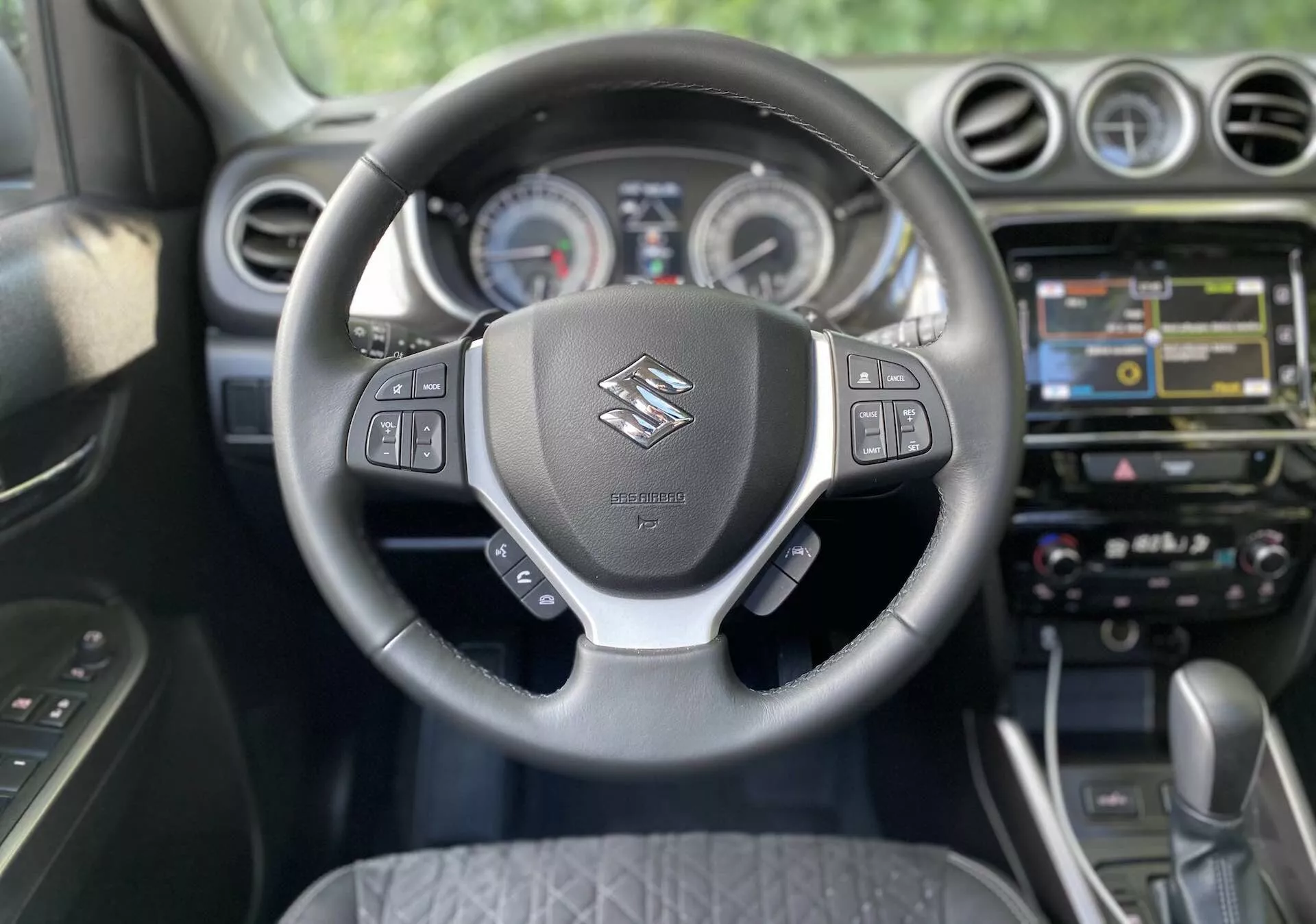 Widok wnętrza Suzuki Vitara 2018 z perspektywy siedzenia kierowcy. Główny nacisk położono na kierownicę z logo Suzuki i wielofunkcyjnymi przyciskami do sterowania audio, poleceniami głosowymi, tempomatem oraz zarządzania wyświetlaczem informacji o pojeździe. Za kierownicą widoczne są analogowe wskaźniki oraz centralny wyświetlacz na desce rozdzielczej. Konsola środkowa zawiera dotykowy system infotainment z nawigacją, multimediami oraz opcjami łączności. Niżej znajdują się dotykowe przyciski i pokrętła do sterowania dwustrefową klimatyzacją. Samochód jest wyposażony w automatyczną skrzynię biegów, co wskazuje na nowoczesny i prawdopodobnie premium projekt kabiny.