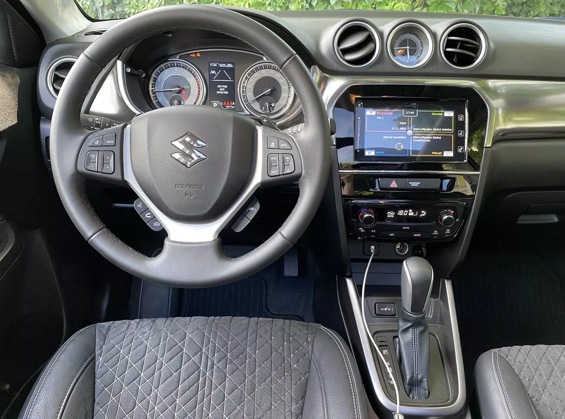 Widok wnętrza Suzuki Vitara 2018 z fotela kierowcy, przedstawiający centralnie umieszczoną kierownicę z logo marki, przyciskami sterowania głośnością, komendami głosowymi i łącznością telefoniczną. Za kierownicą znajduje się cyfrowo-analogowy zestaw wskaźników z prędkościomierzem, obrotomierzem i centralnym wyświetlaczem cyfrowym. Deska rozdzielcza wyposażona jest w dotykowy ekran infotainment z nawigacją GPS, radio i system klimatyzacji. W widoku wyróżniają się automatyczna dźwignia zmiany biegów i elektroniczny hamulec postojowy, wraz z pikowanym designem fotela i okrągłymi otworami wentylacyjnymi, oddając nowoczesną technologię samochodową i ergonomiczną konstrukcję.