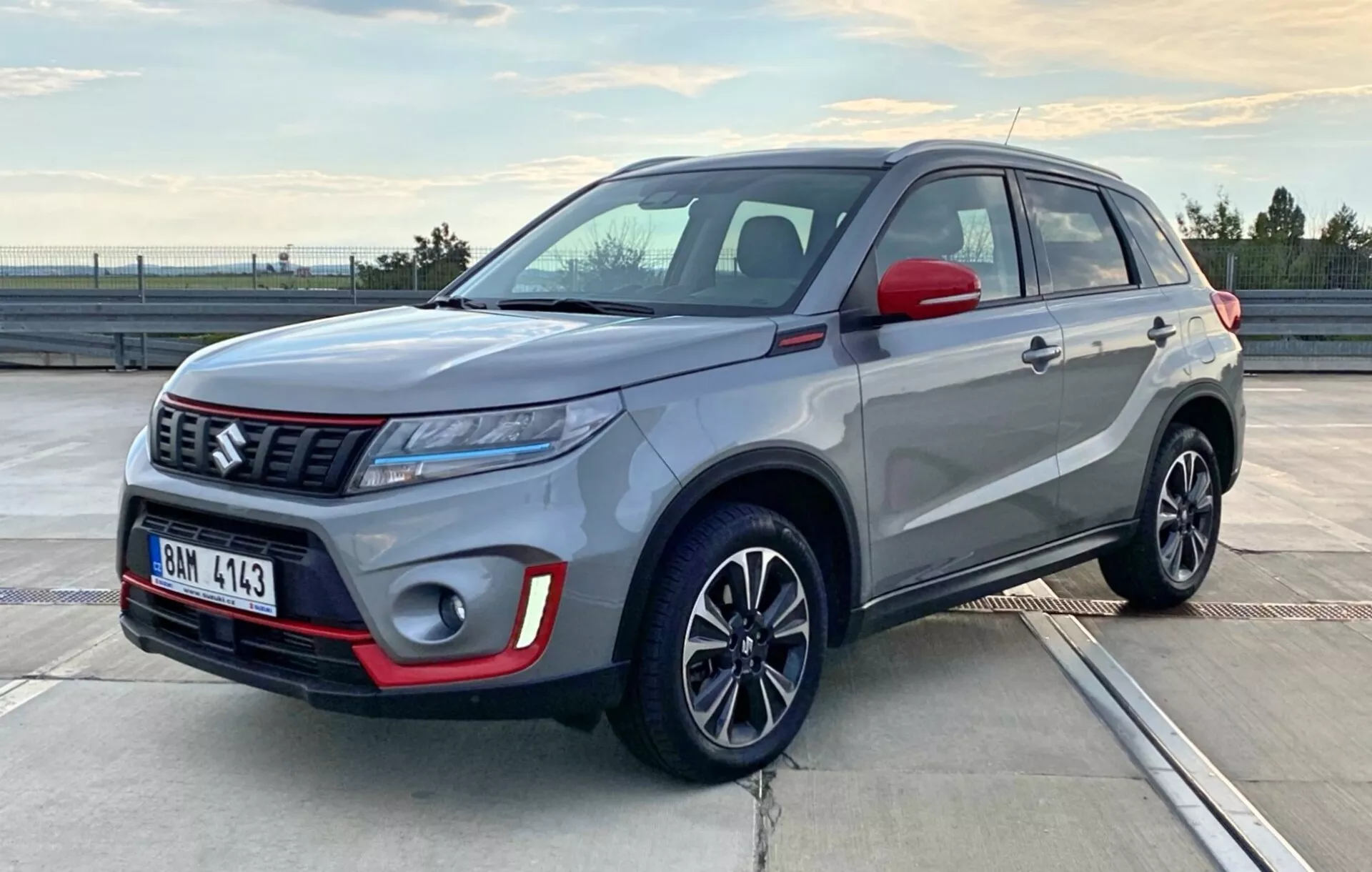 Obraz przedstawia kompaktowy SUV Suzuki Vitara 2018 zaparkowany na parkingu na otwartym terenie podczas wschodu lub zachodu słońca. Fotografia uchwyciła samochód z przodu pod kątem trzech czwartych, ujawniając stronę kierowcy i przód, podkreślając konfigurację reflektorów, styl grilla, przedni zderzak, koła i boczne linie nadwozia. Pojazd ma dwukolorową karoserię: srebrno-szary nadwozie i czarny dach oraz lusterka boczne, z czerwonymi akcentami na przednim zderzaku. Widać również eleganckie reflektory LED, wyraźny emblemat marki na wzorzystej kratce oraz wieloramienne felgi ze stopu, co pokazuje współczesny i sportowy design pojazdu.