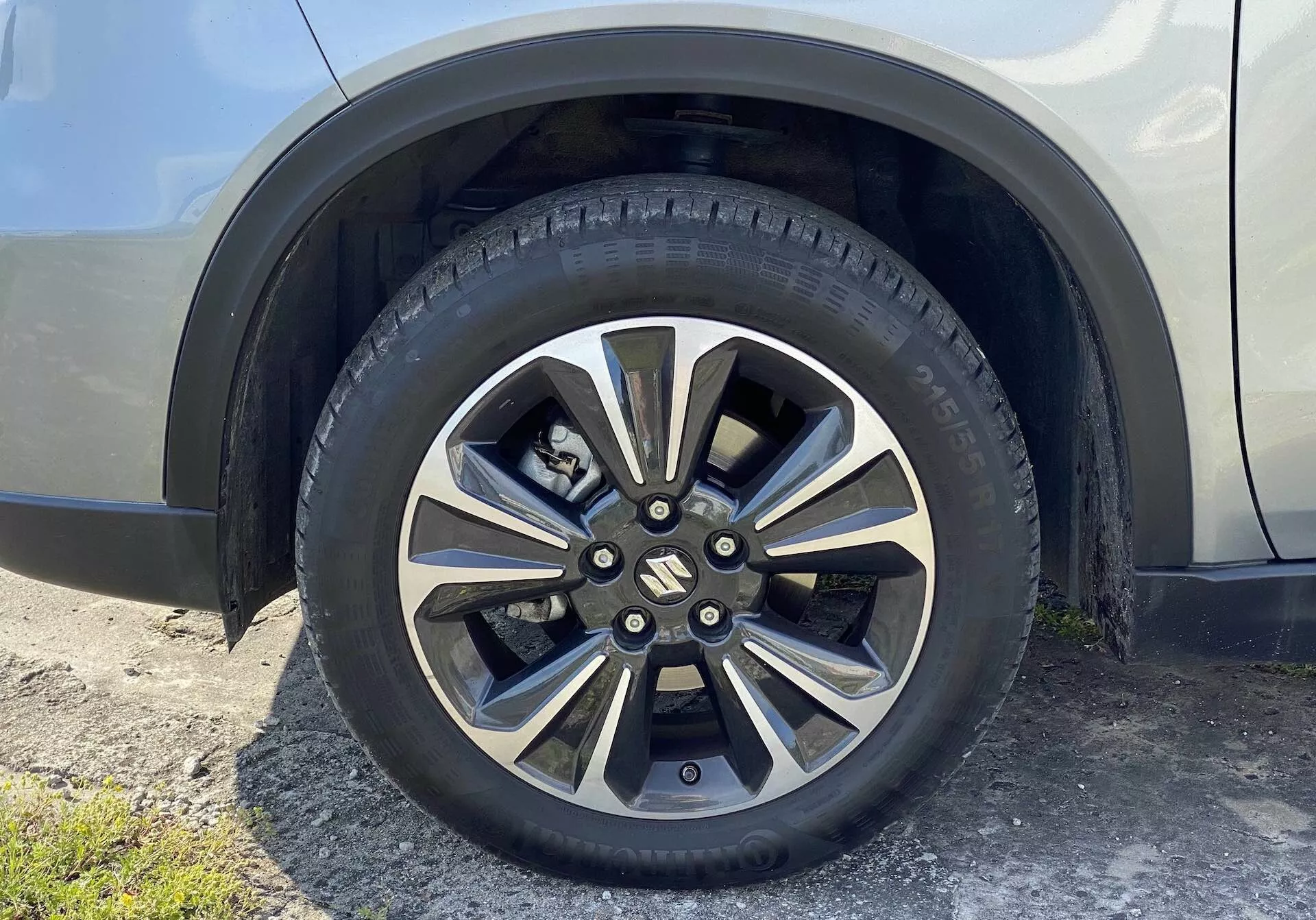 Zbliżenie na koło Suzuki Vitara 2018, pokazujące aluminiowy wzór z wieloma szprychami w metalicznym i ciemniejszym malowanym wykończeniu. Markowy centralny dekiel zakrywa nakrętki i piastę, a opona na drogę ma widoczne wzory bieżnika oraz oznaczenia boczne. W tle widoczna jest część błotnika i panelu bocznego samochodu w neutralnym, połyskliwym kolorze, widziana z lekko wznoszącej się perspektywy z poziomu ziemi.