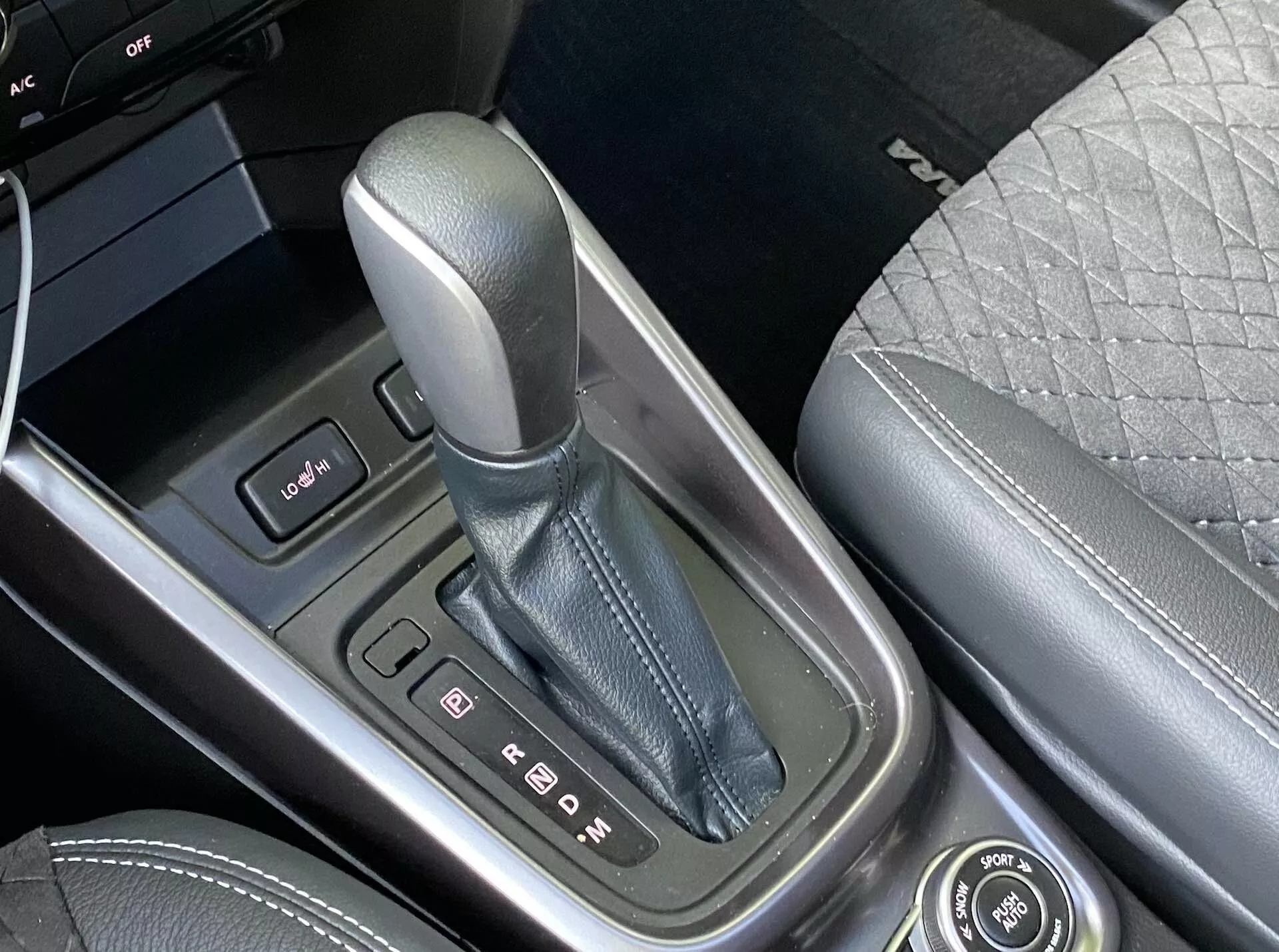 Zbliżenie środkowej konsoli Suzuki Vitara 2018, z naciskiem na dźwignię zmiany biegów w pozycji 'P' (parkowanie), otoczoną metalicznym wykończeniem z wyraźnymi wskaźnikami biegów. Po lewej stronie widoczny jest panel kontrolny z dwoma przyciskami oraz krawędź ekranu. Środkowa konsola wykonana jest z wysokiej jakości materiałów, a po prawej stronie można dostrzec część przedniego siedzenia z pikowanym wzorem. Obecne jest również pokrętło do funkcji infotainment, co podkreśla zaawansowaną technologię i luksusowe cechy pojazdu.
