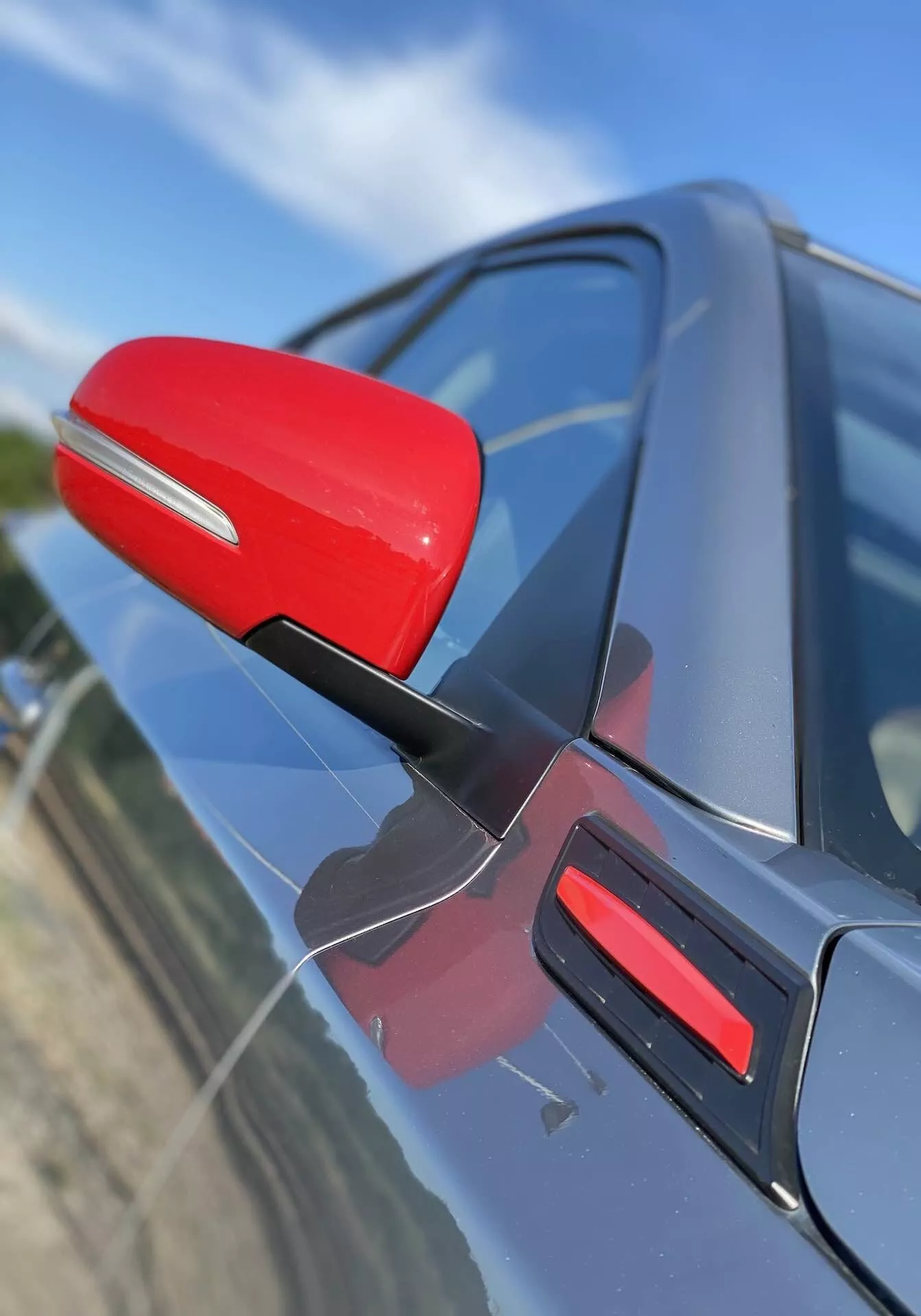 Zbliżenie lusterka bocznego po stronie kierowcy w Suzuki Vitara 2018, wyróżnionego czerwonym kolorem na tle ciemnej karoserii samochodu. Lusterko jest przymocowane na smukłym, czarnym wsporniku, co sugeruje aerodynamiczną wydajność. Ujęcie uchwyca lusterko pod lekko przednim kątem skośnym, z perspektywą skierowaną w dół, z pasującym czerwonym wykończeniem na drzwiach. Tło jest rozmyte, podkreślając detale lusterka i designu drzwi samochodu.