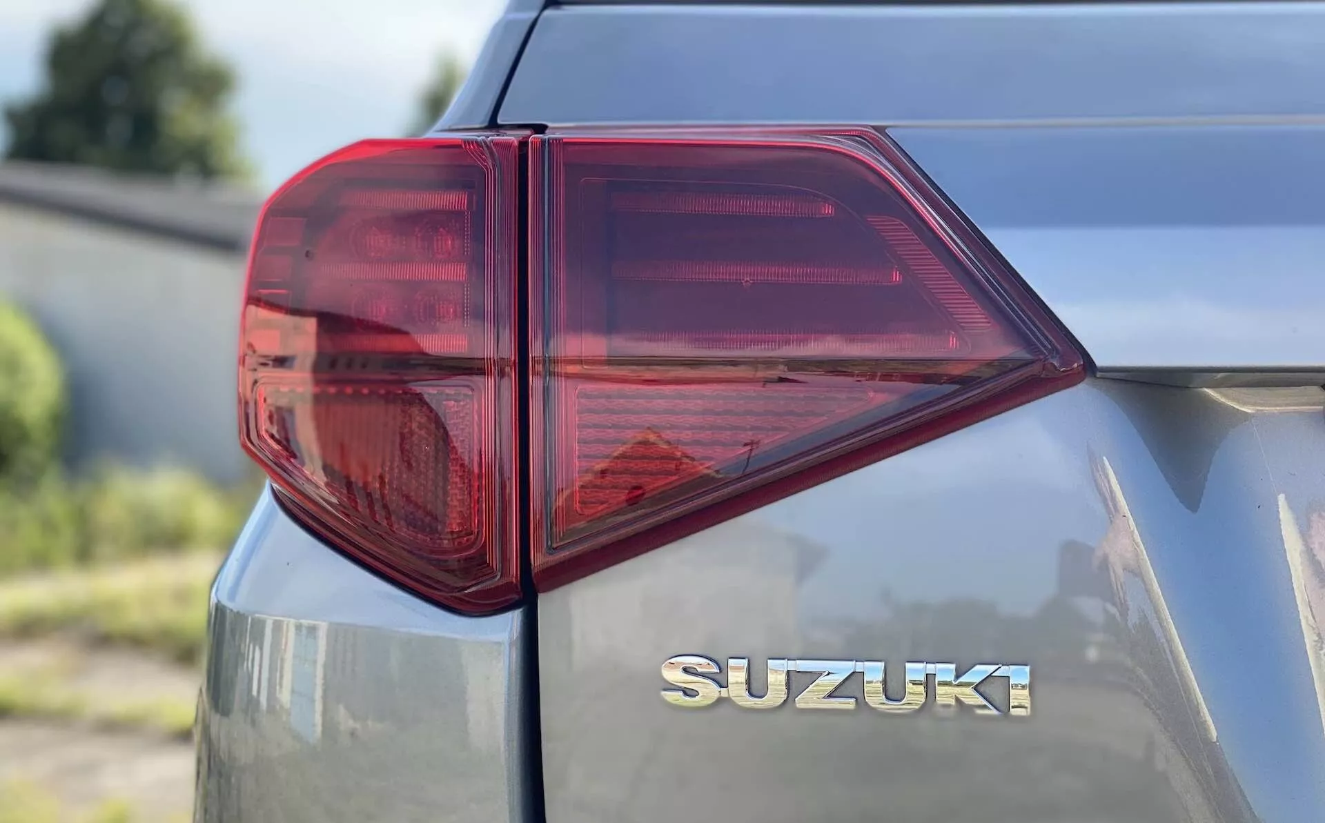 Zbliżenie widoku tylnej lewej strony Suzuki Vitara 2018 z fokus na tylne światło. Perspektywa w trzech czwartych od boku, ujęta z niskiej lub średniej wysokości, ukazuje charakterystycznie stylizowane tylne światło LED z ostrymi kątami i kilkoma komorami dla różnych funkcji, takich jak światło hamowania, kierunkowskaz i możliwie światło przeciwmgielne. Przewaga czerwonego z przezroczystą sekcją prawdopodobnie dla świateł cofania, podkreśla nowoczesną technologię oświetleniową motoryzacji. Tylny bagażnik wyposażony jest w chromowany znaczek 'SUZUKI' umieszczony centralnie pod tylną szybą, co podkreśla czyste i gładkie oznaczenie marki. Metalizowane wykończenie odbija nieco światła, wskazując na dobrze utrzymaną lub nowszą powierzchnię pojazdu.