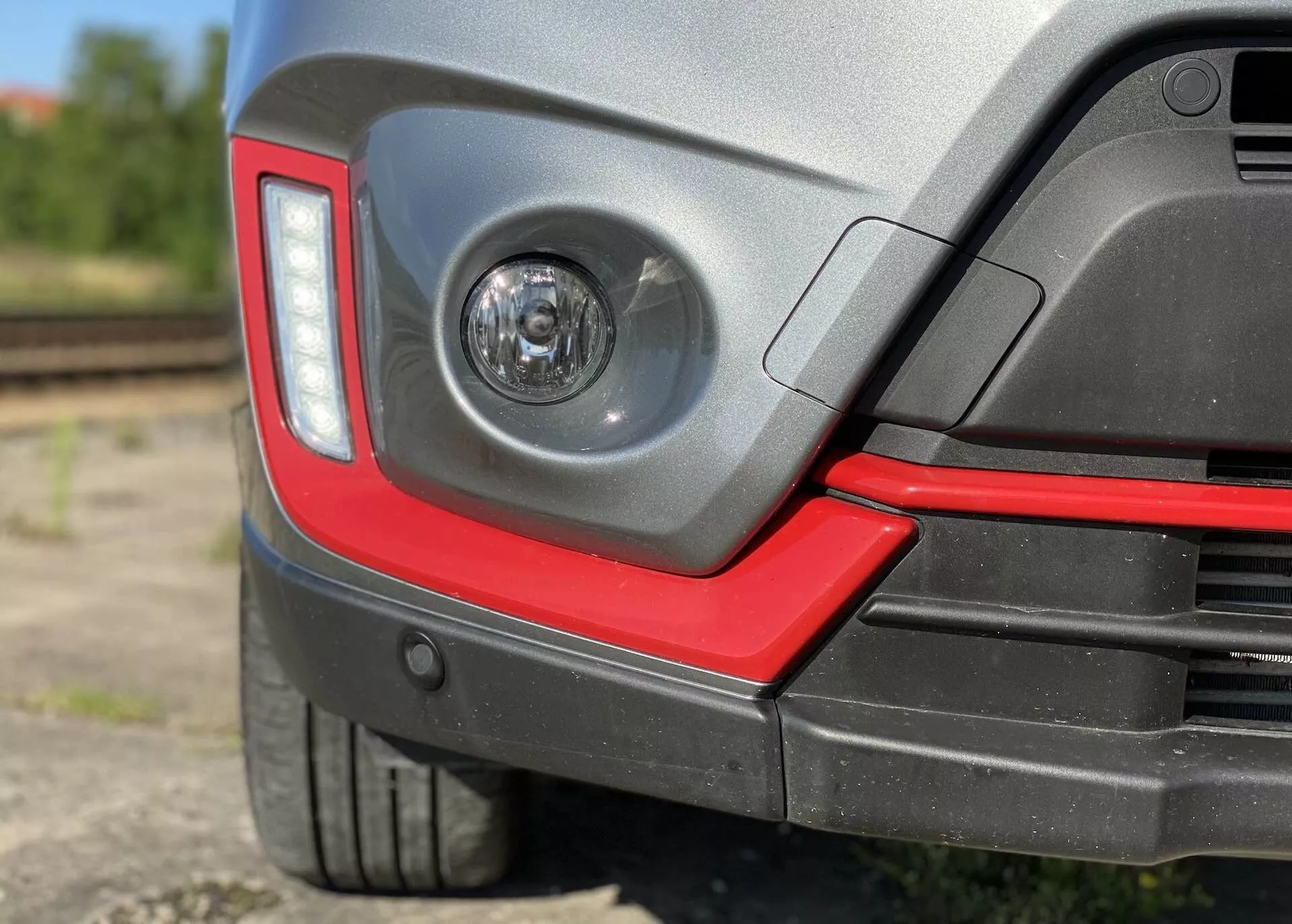 Zbliżenie tylnej części samochodu Suzuki Vitara 2018, z wyraźnym światłem przeciwmgielnym w tylnym zderzaku, nowoczesnym kątowym kształtem tylnych świateł, dwukolorowym tylnym zderzakiem ze sportowym czerwonym akcentem, potencjalnymi czujnikami parkowania, niskoprofilową oponą z plastikowym nadkolem ochronnym, błyszczącym wykończeniem lakieru i rozmytym półprzemysłowym tłem z torami kolejowymi.