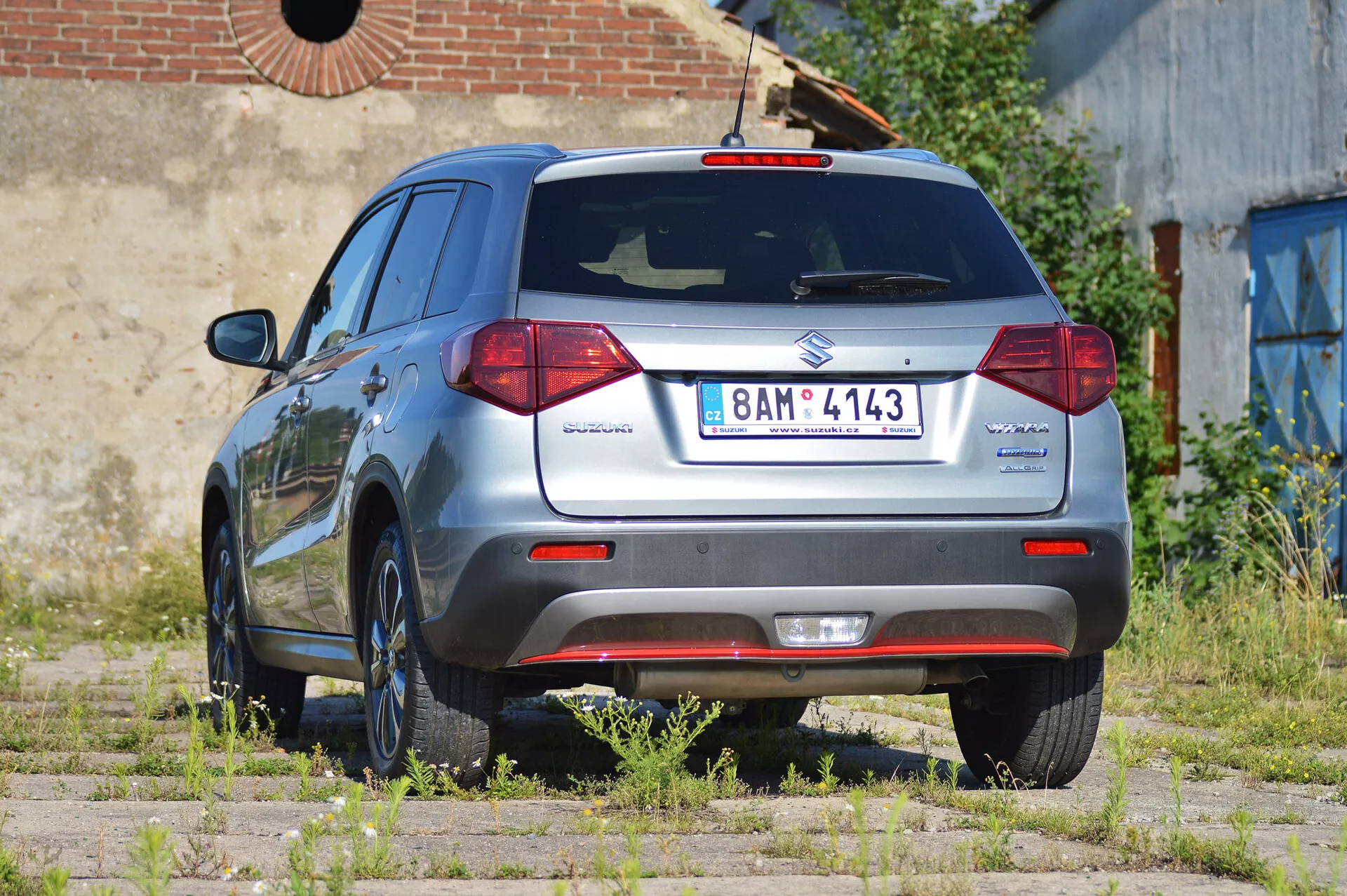 Widok tylno-boczny na metalizowany szary samochód hatchback Suzuki Vitara 2018 uchwycony w świetle dziennym. Obraz podkreśla design tyłu samochodu, z wyraźnymi tylnymi światłami, wycieraczką tylnej szyby i emblematami Suzuki i Vitara. Pokazuje również odznakę 'Allgrip' oznaczającą napęd na cztery koła, europejską tablicę rejestracyjną, aluminiowe felgi, czujniki parkowania na tylnym zderzaku i antenę zamontowaną na dachu. Pojazd jest zaparkowany na nierównym terenie z trawą i ziemią, co sugeruje zewnętrzny, ewentualnie zaniedbany obszar. Samochód jest czysty i zadbany.
