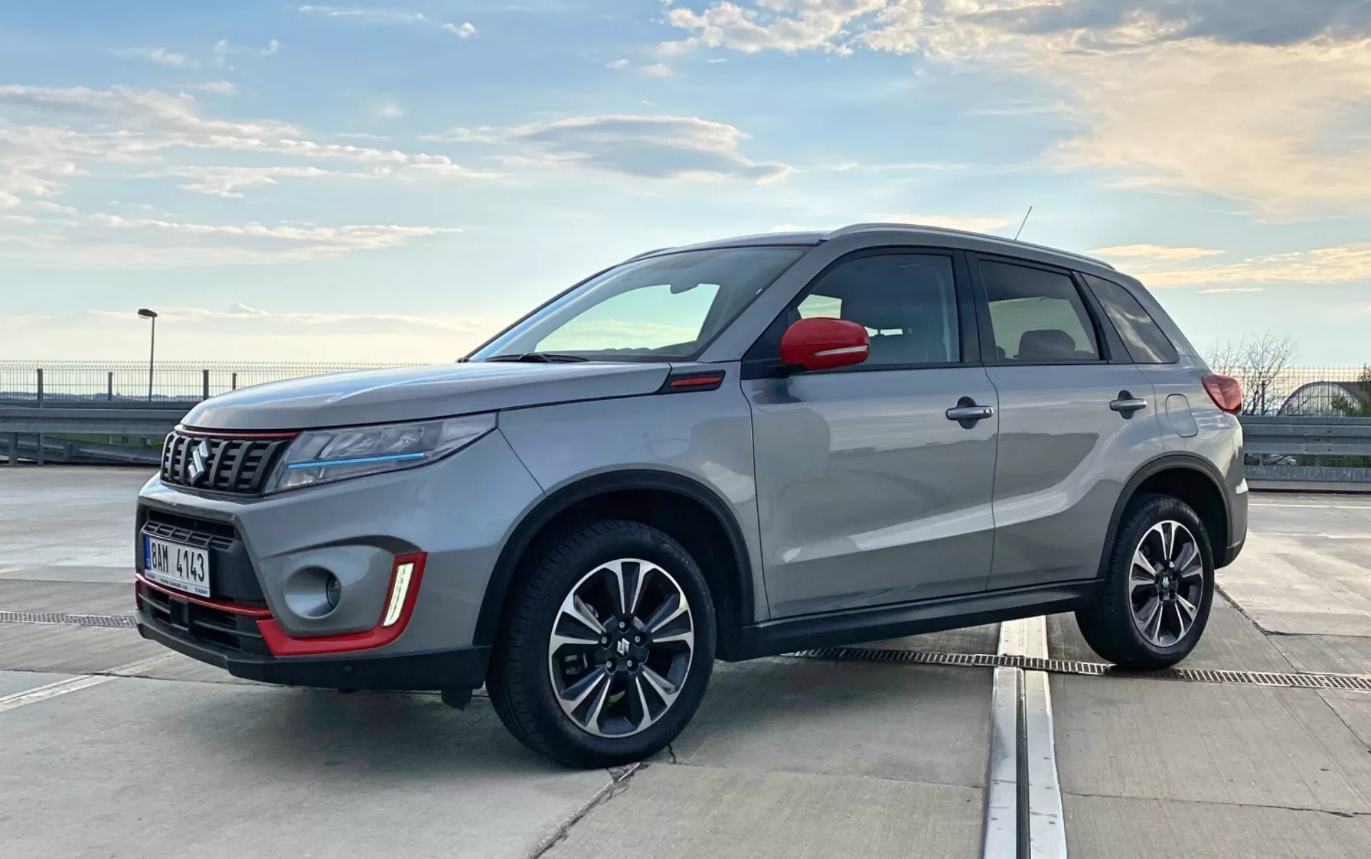 Obraz przedstawia Suzuki Vitara 2018, nowoczesne kompaktowe SUV, zaparkowane na zewnątrz w świetle dziennym. Jest uchwycone z widoku trzy-czwartowego z przodu, podkreślając zarówno przedni, jak i boczny profil. Kąt jest nieco niski, na poziomie klamek drzwi, co sugeruje, że fotograf kucał lub się pochylał. Widoczne szczegóły obejmują przedni grill, reflektory, obudowę lamp przeciwmgielnych, emblemat, lusterka boczne, felgi aluminiowe z zintegrowanymi kierunkowskazami, klamki drzwi, listwy okienne, linię dachu i relingi dachowe. Reflektory projekcyjne i elegancka antena sugerują zaawansowane technologie. Nowoczesne elementy projektowe SUV wskazują na funkcje takie jak światła LED i możliwy bezkluczykowy system wejścia.
