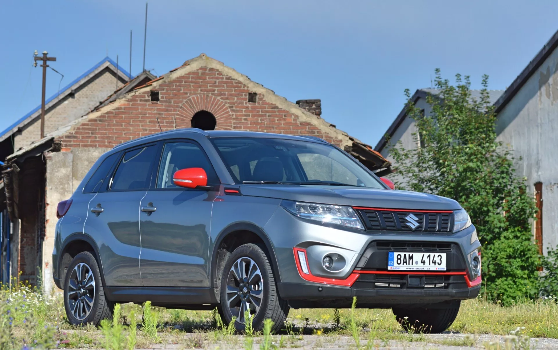 Obraz przedstawia SUV Suzuki Vitara 2018 z niskiej perspektywy przedniego widoku z trzech czwartych, podkreślając przednią prawą stronę pojazdu z zauważalnym prześwitem. Widoczne cechy obejmują wytrzymały przedni zderzak ze światłami przeciwmgielnymi i srebrną płytą poślizgową, emblemat marki na poziomej kratce, wyraźnie zarysowane linie maski, nowoczesne reflektory prawdopodobnie z technologią LED lub HID, wieloramienne felgi aluminiowe, czerwone lusterka boczne z wbudowanymi kierunkowskazami, muskularne błotniki, odblaskową szybę przednią i przyciemniane okna. Vitara jest zaparkowana na nieutwardzonej trawiastej powierzchni, przed starym budynkiem dodającym przemysłowe lub wiejskie tło, sugerującym przydatność w środowiskach zewnętrznych.