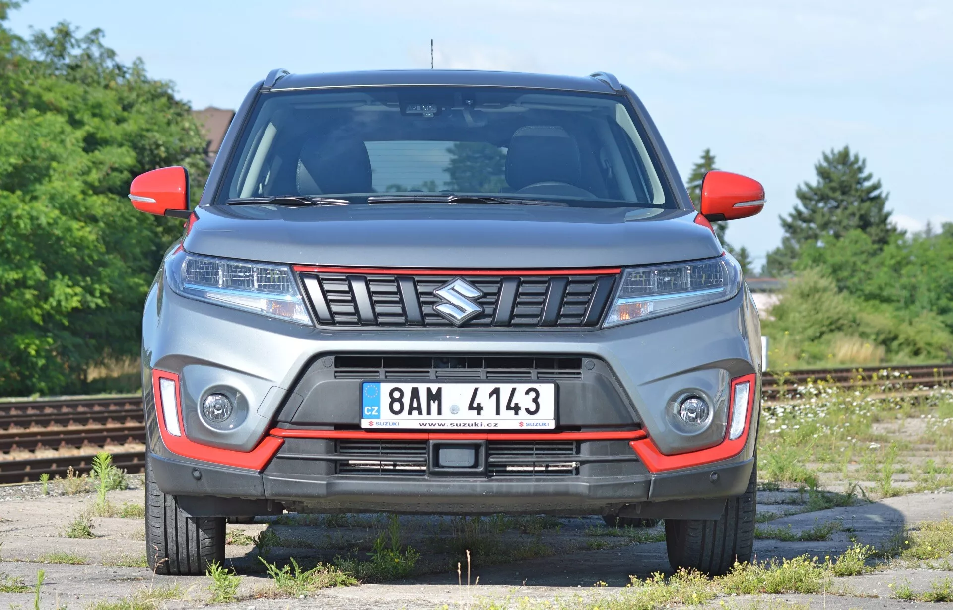 Widok od przodu Suzuki Vitara 2018 z lekko niskiego kąta. Obraz podkreśla cechy przedniej części pojazdu, w tym maskę, kratkę (z emblematem Suzuki), eleganckie reflektory (potencjalnie LED) i dodatkowe oświetlenie zderzaka. Samochód wygląda na solidny, co sugeruje możliwości terenowe. Widoczne są kierunkowskazy w lusterkach bocznych i przednia tablica rejestracyjna. Szara karoseria z czerwonymi akcentami może oznaczać specjalny zestaw wykończeniowy. Tło wydaje się być opuszczoną strefą przemysłową lub placem kolejowym, co zapewnia wyraźny widok na design samochodu.